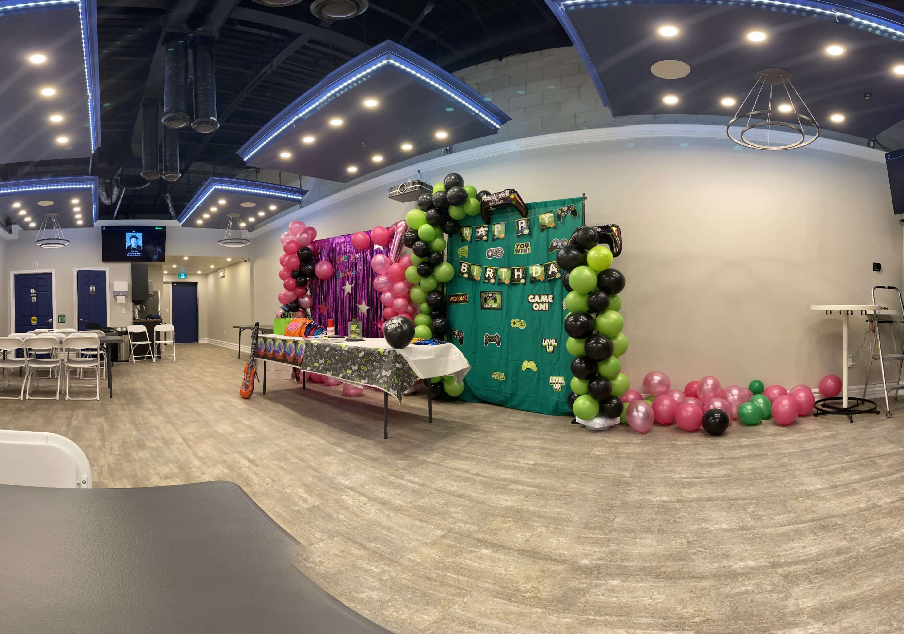 A party room decorated with a vibrant backdrop, an array of balloons in black, green, and pink, and tables set up for guests.
