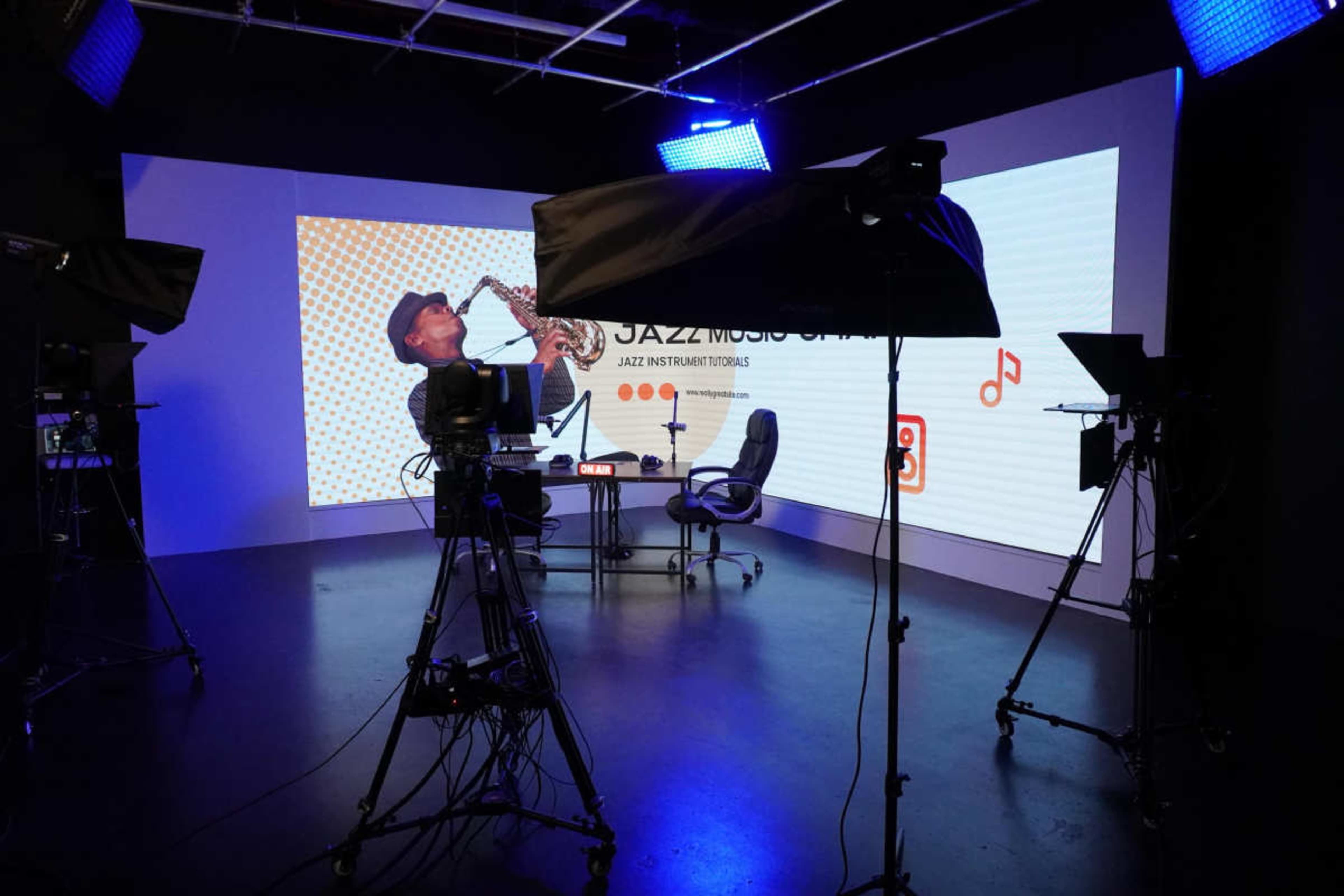 A film studio is equipped with cameras and lighting, featuring a large screen displaying a jazz musician playing the saxophone.