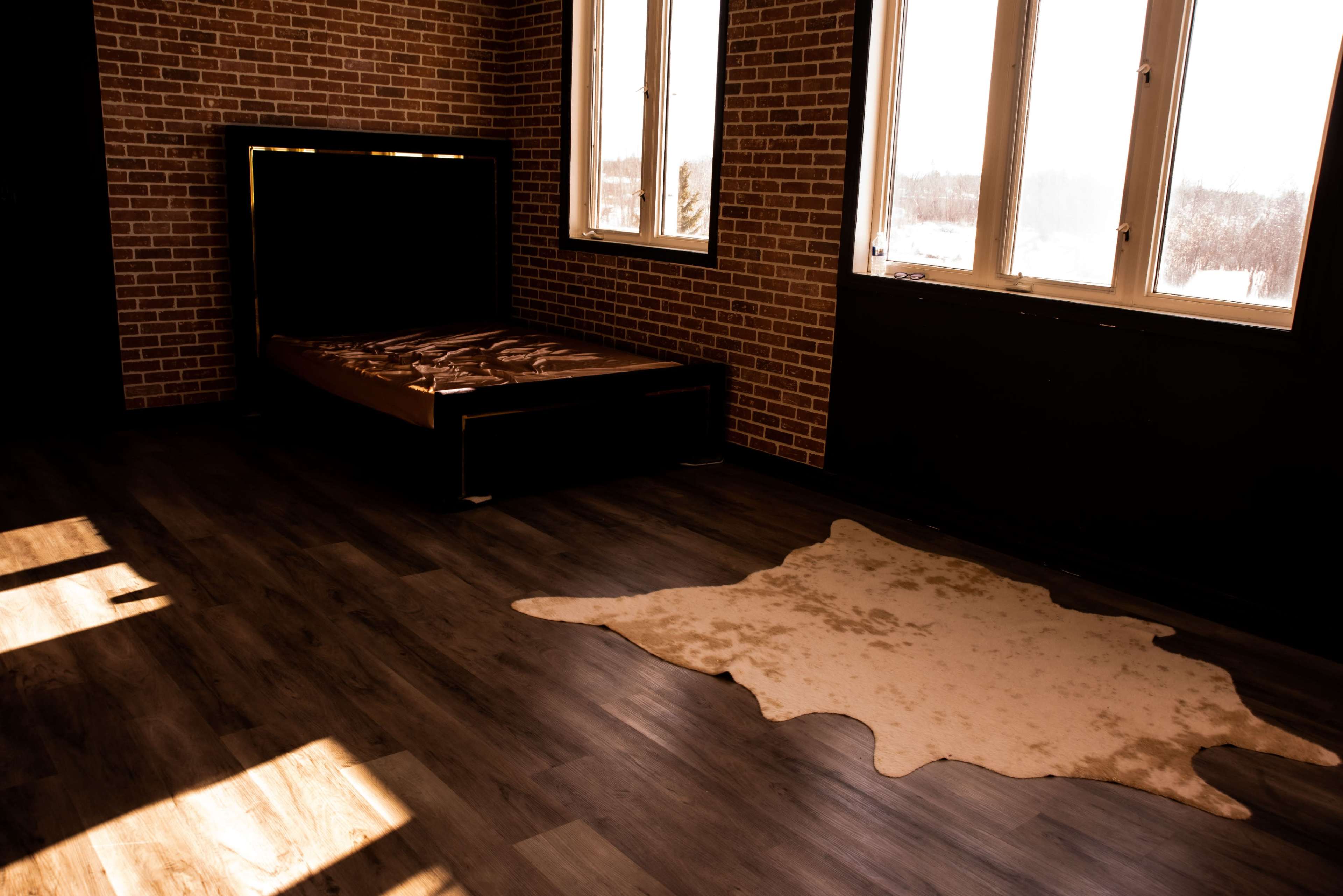 The interior of a minimalist bedroom features a bed against a brick wall and a light-colored rug on the wooden floor.