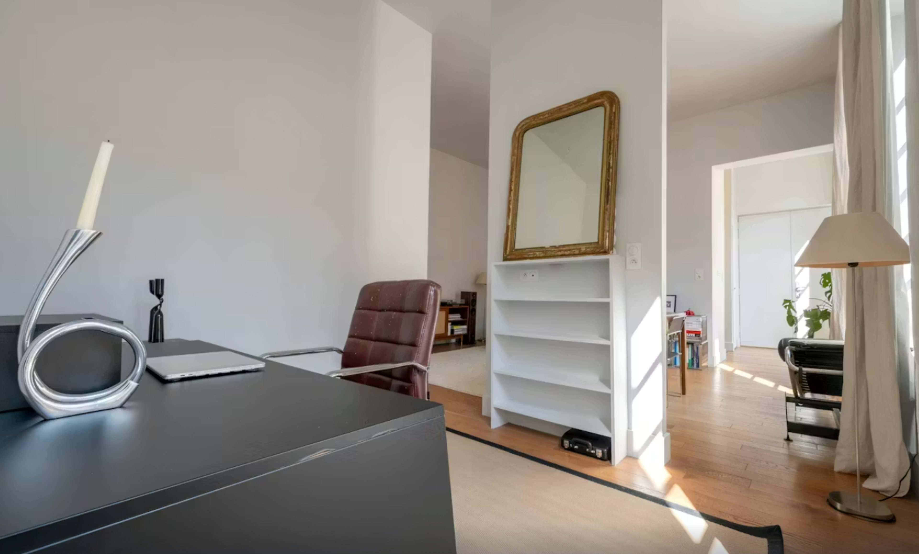 A modern office space features a dark desk with a laptop, a red leather chair, a decorative mirror on a white wall, and an open doorway leading to a bright area.
