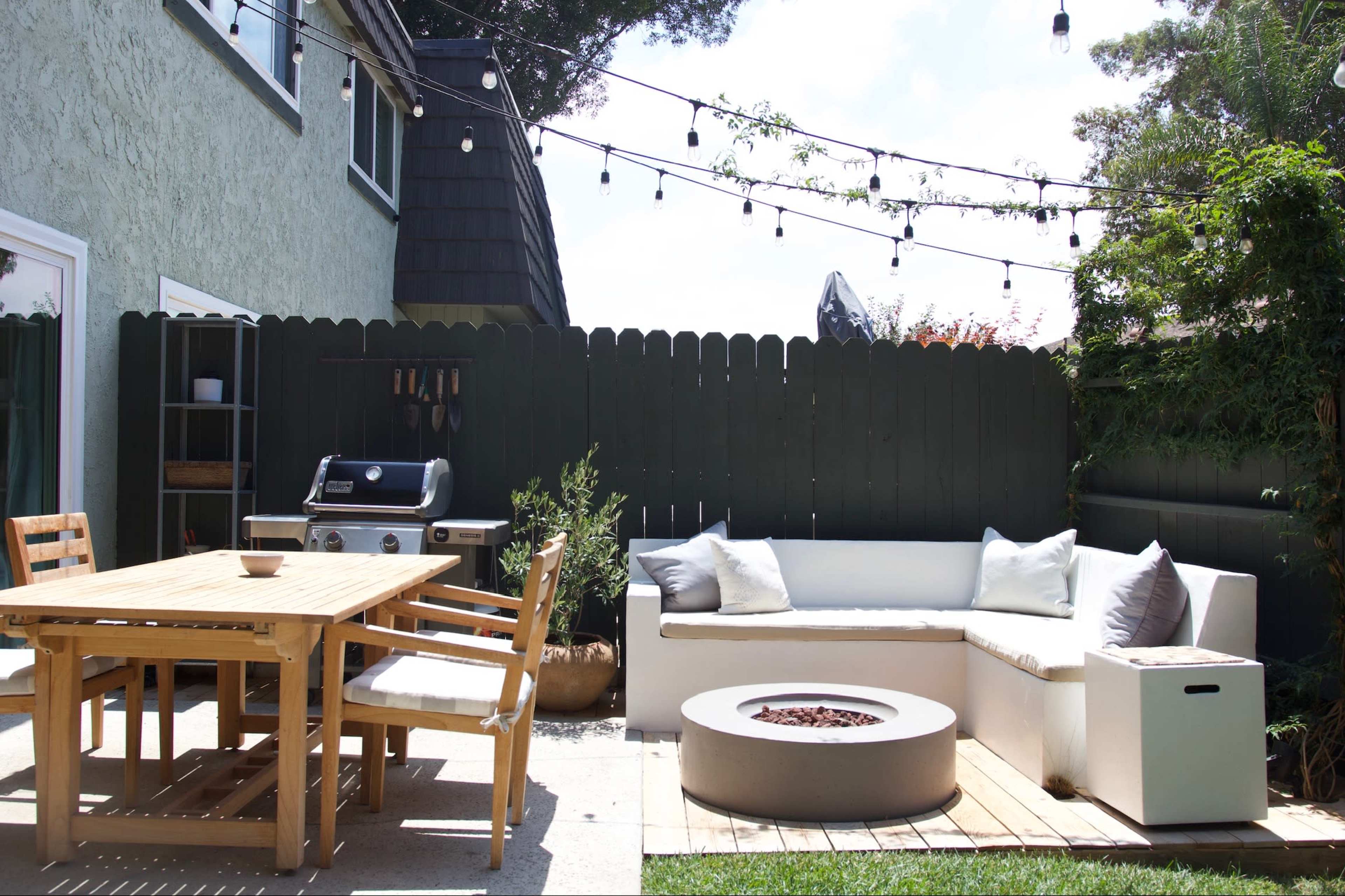 The image shows a backyard patio featuring a wooden dining table with chairs, a sectional sofa, a fire pit, a grill, and string lights overhead.
