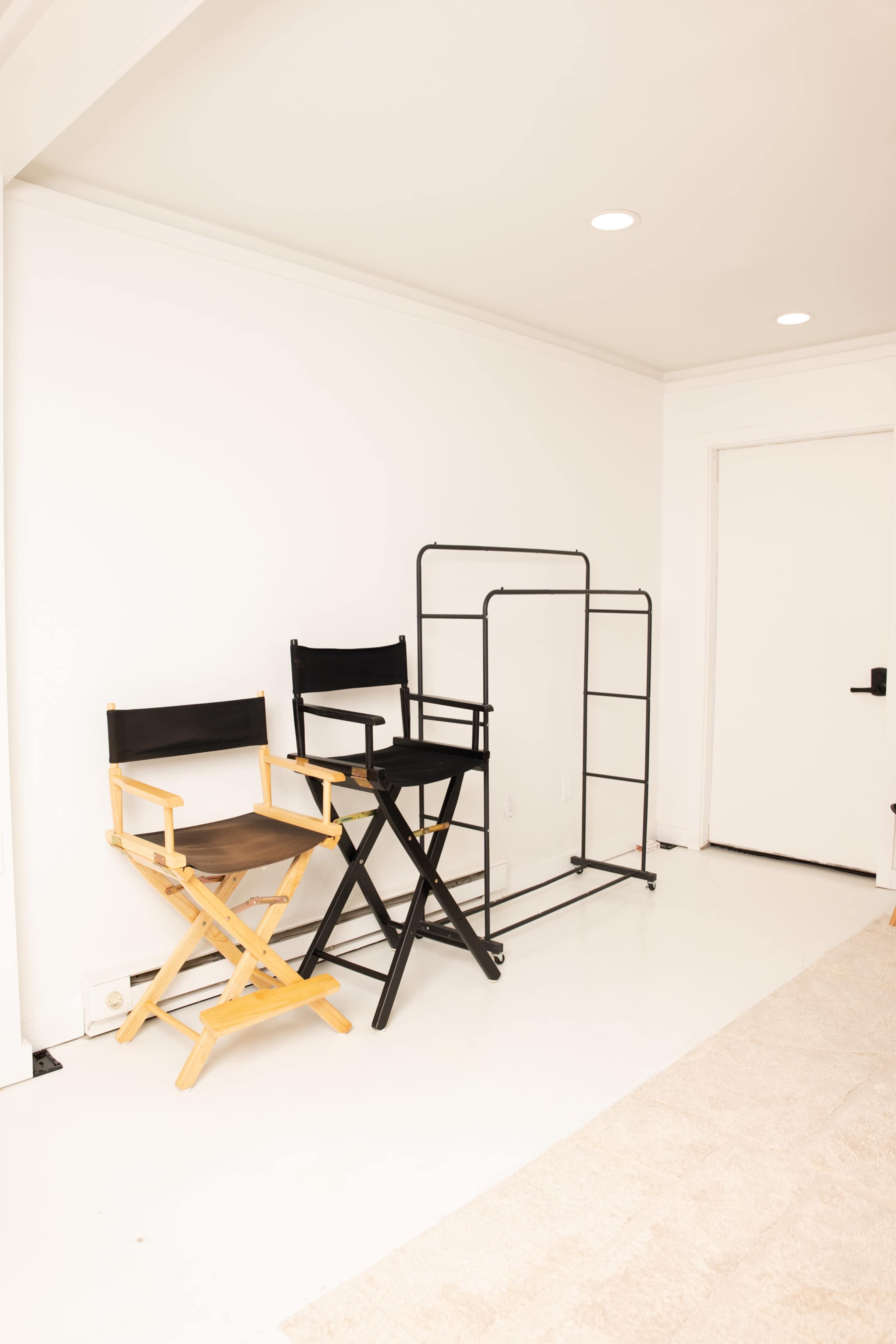 The image shows two director's chairs, one with a wooden frame and the other with a black frame, positioned beside a clothing rack against a white wall.