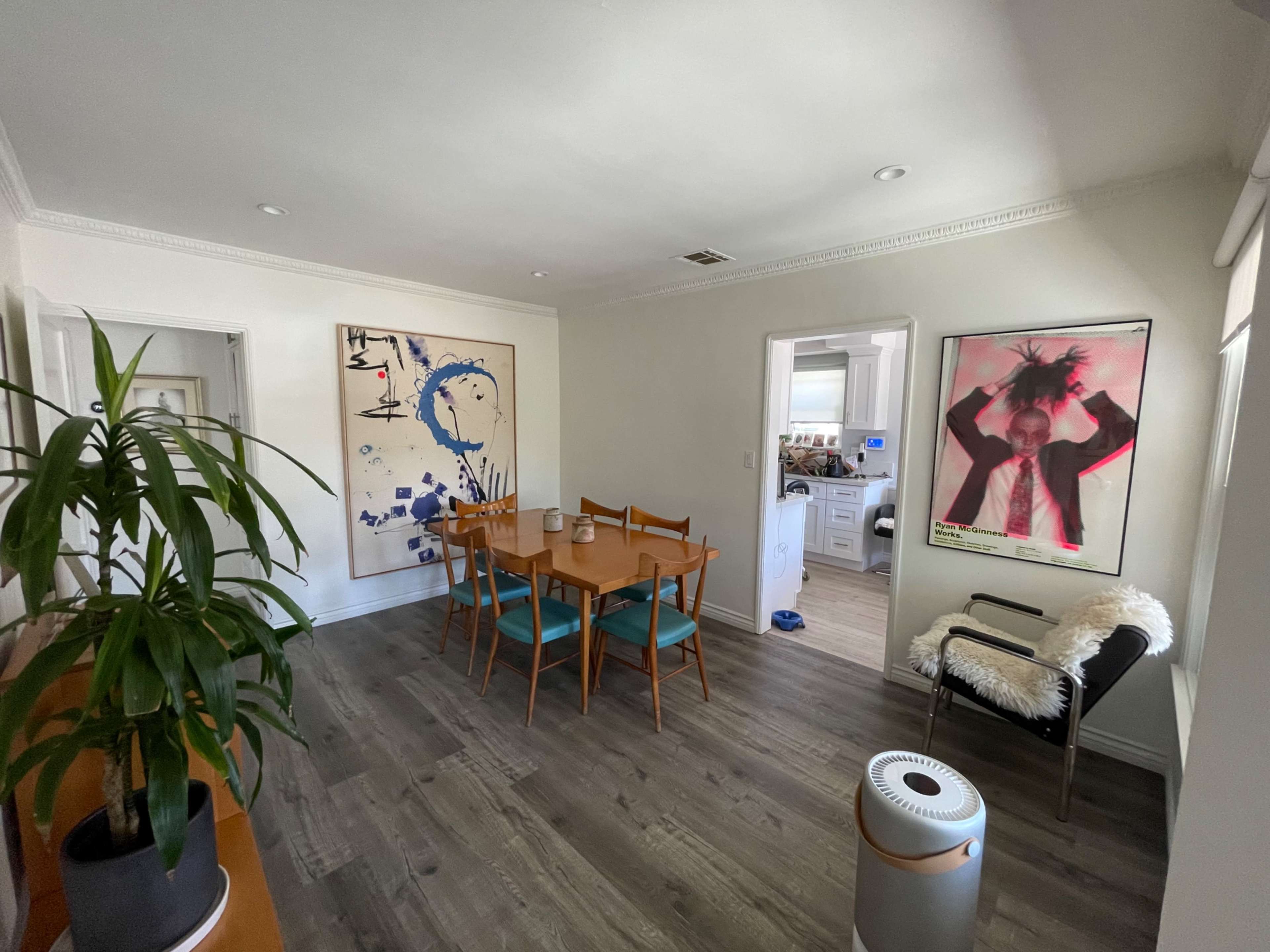 A dining area features a wooden table surrounded by chairs, with large artwork on the walls and a plant in the corner.