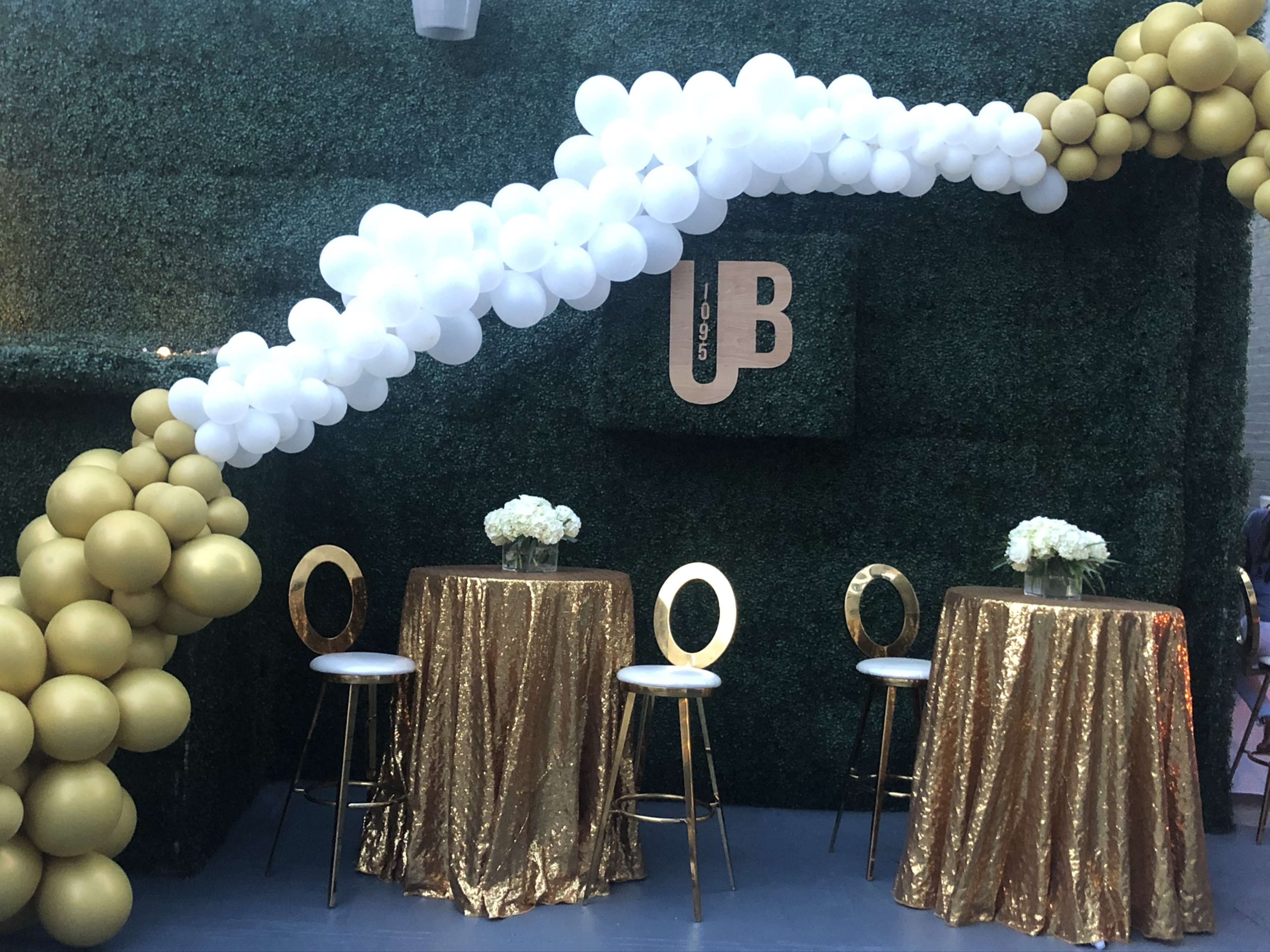 A decorated area with two round tables draped in gold sequined tablecloths, surrounded by an arch of white and gold balloons against a green backdrop.