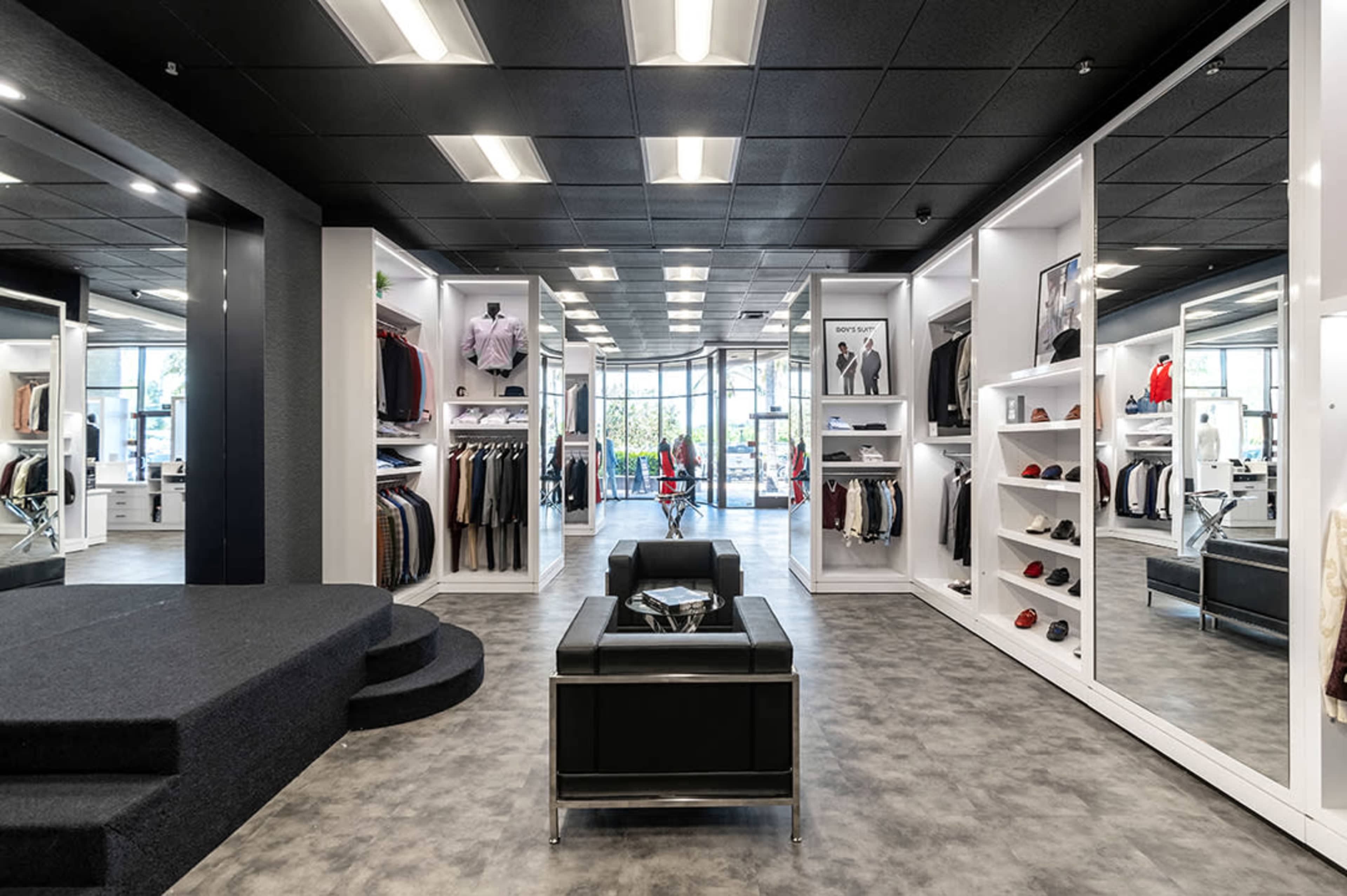 A modern clothing store featuring a spacious layout with clothing displays arranged on white shelves, mirrors lining the walls, and a seating area in the center.
