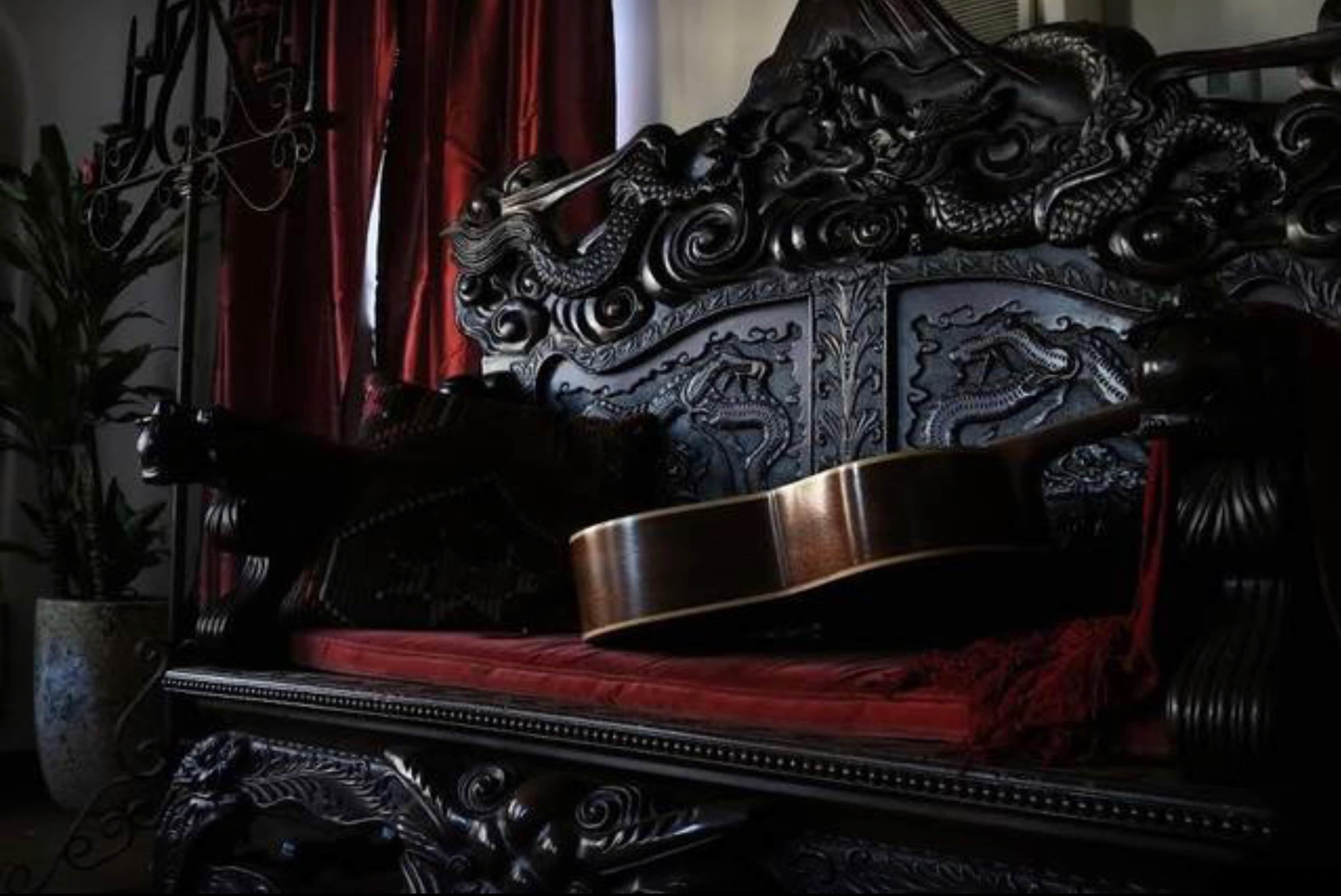 A detailed, dark wooden bench with intricate carvings is adorned with a red fabric cushion and features an acoustic guitar resting against it.