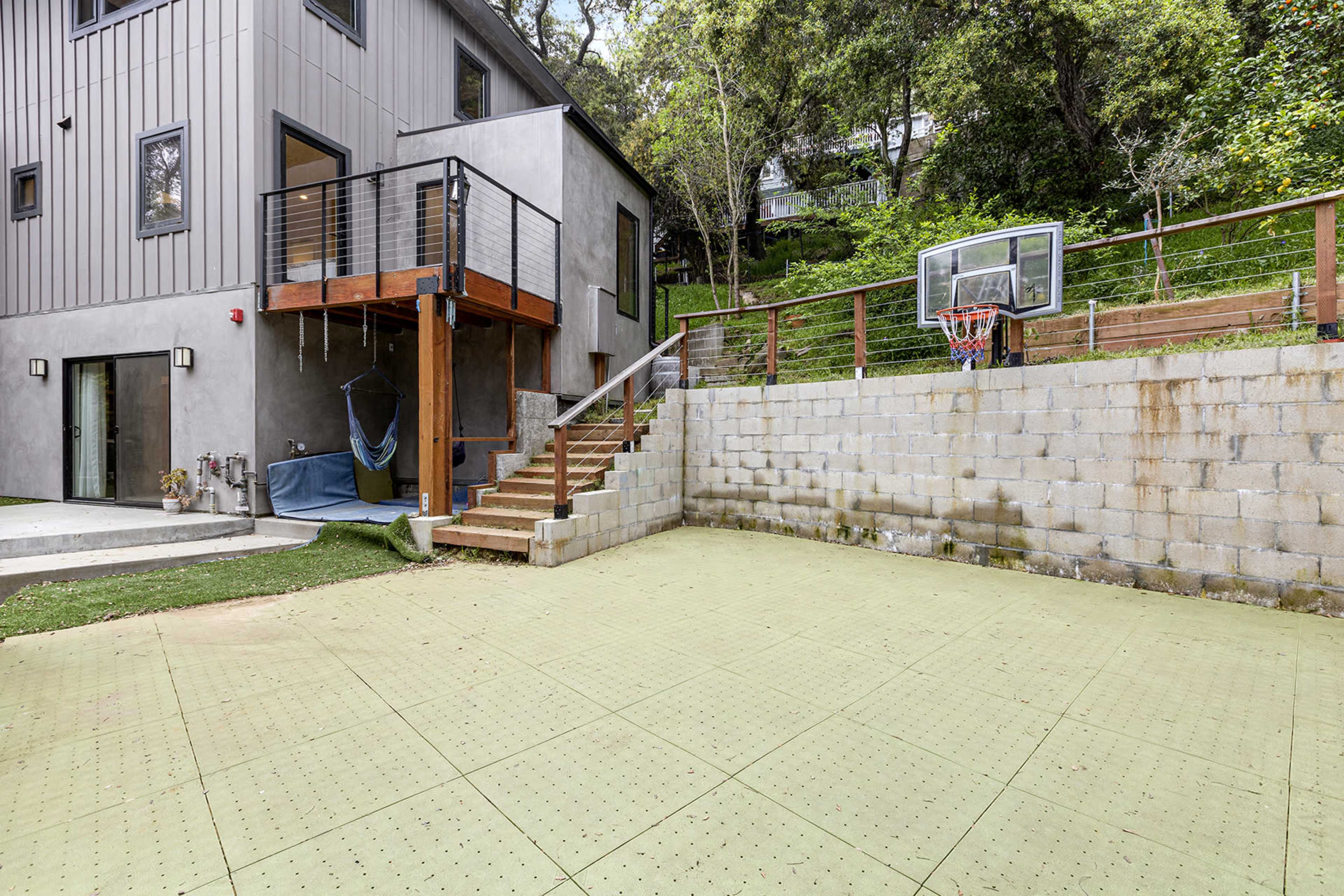 The image shows a backyard area with a concrete basketball court next to a modern house featuring a wooden staircase and a hammock.