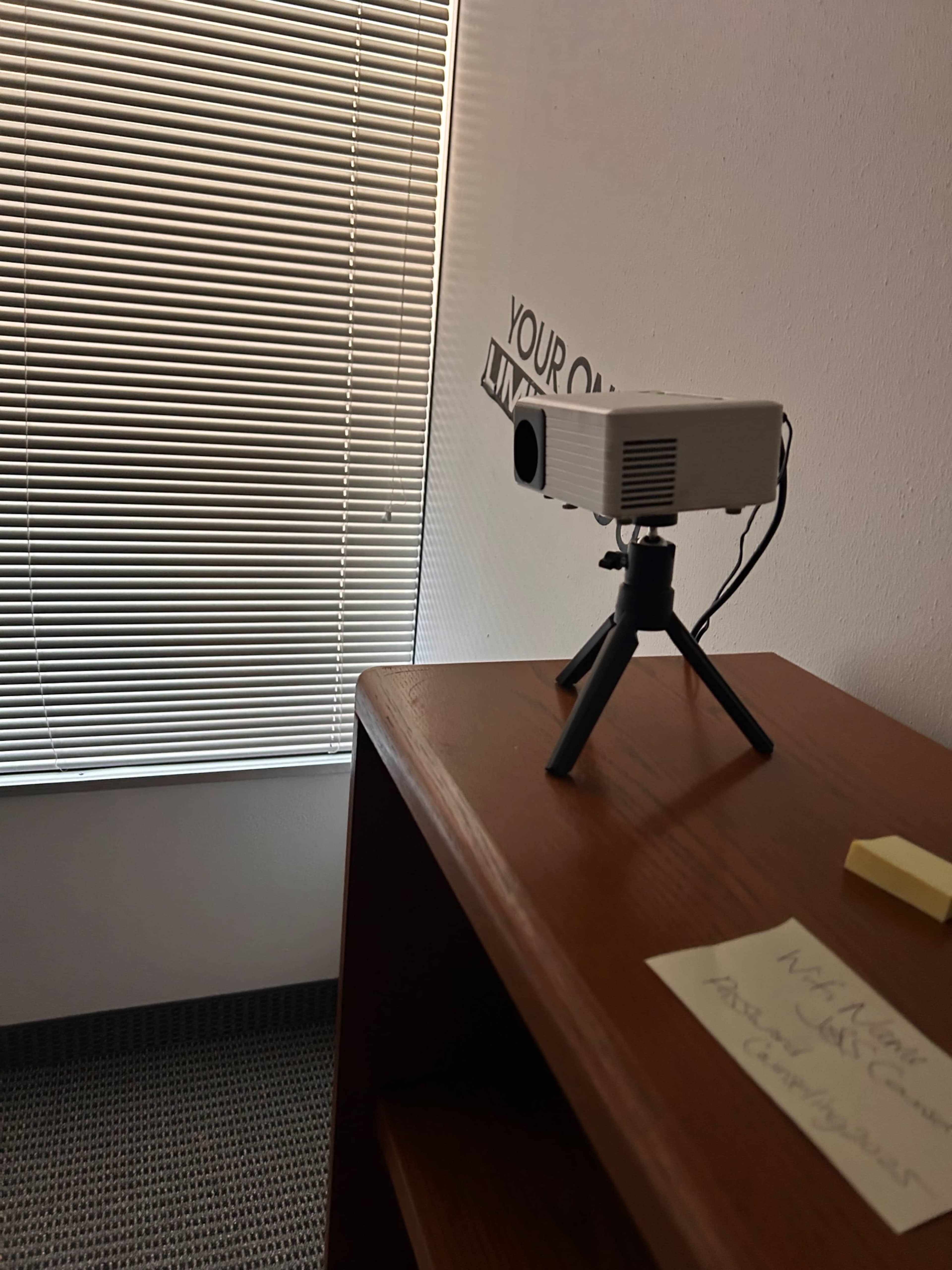 A projector is positioned on a small tripod atop a wooden shelf, with a partially closed blind casting shadows in the background.