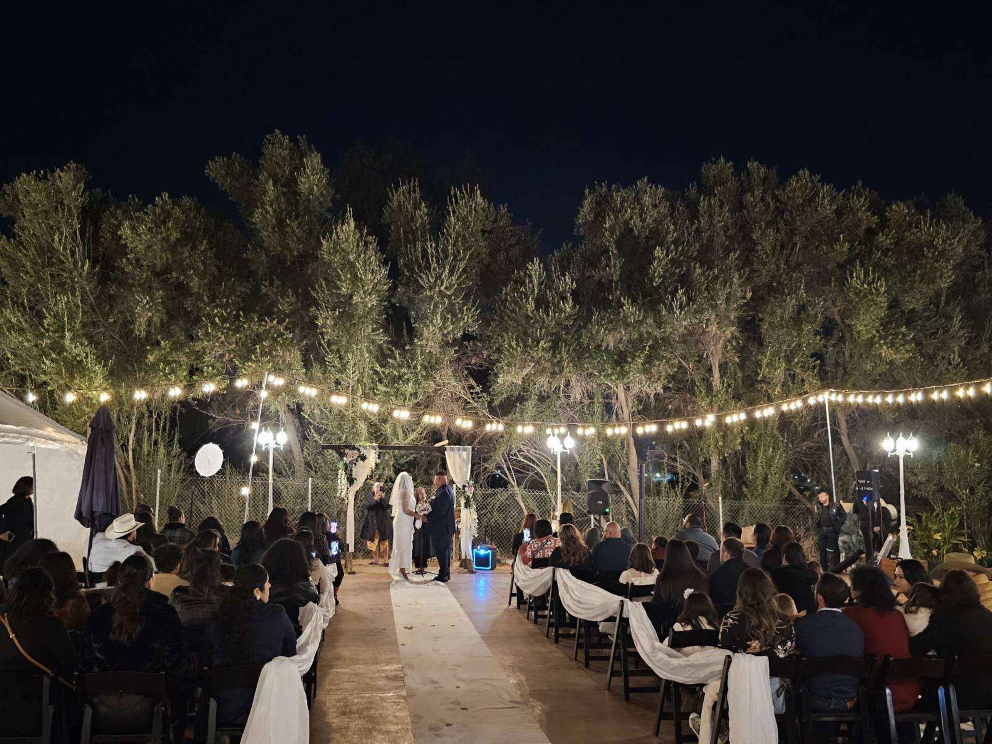 A couple exchanges vows on an outdoor stage illuminated by string lights, surrounded by seated guests in an evening setting.