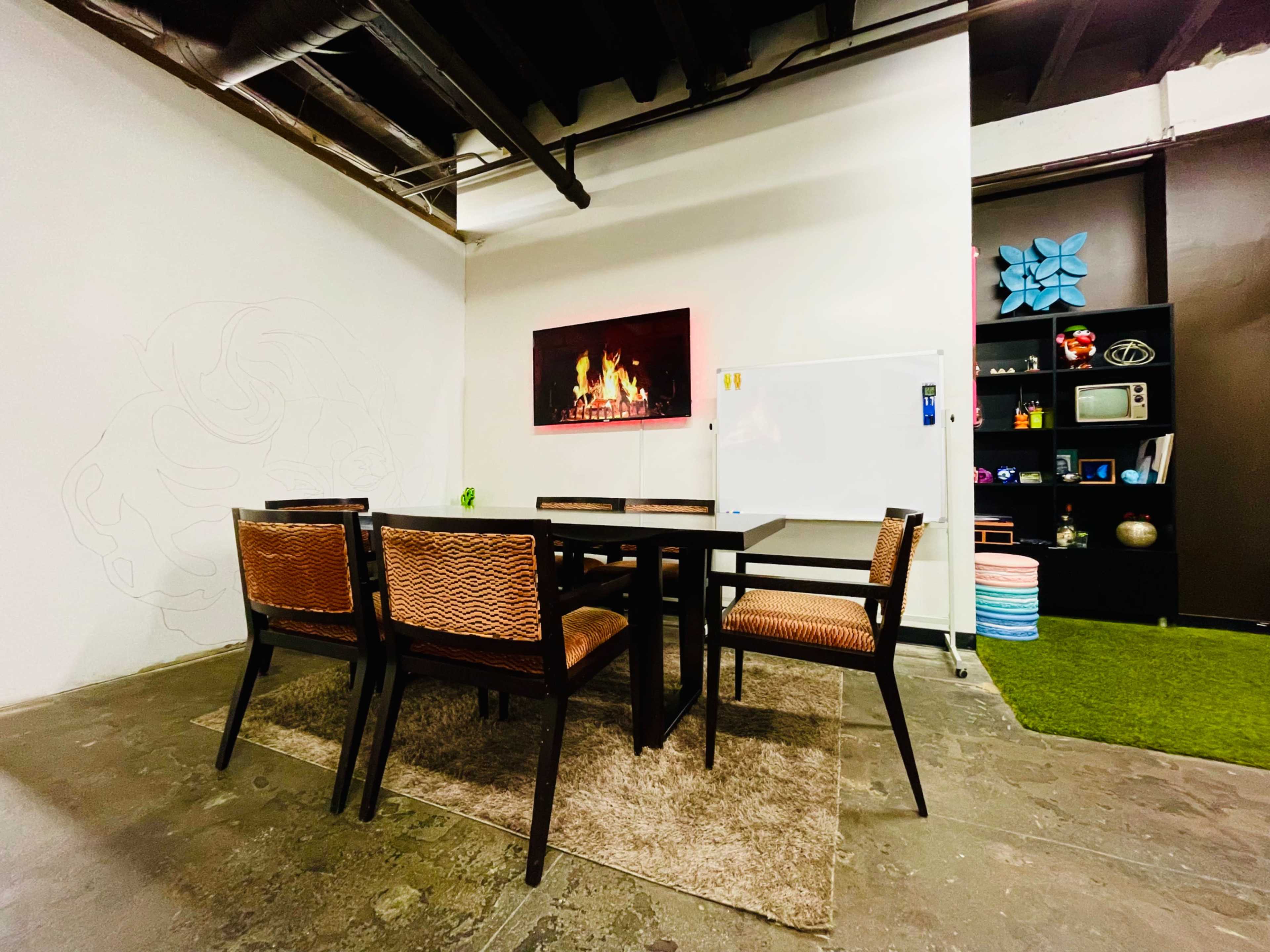 A conference room features a rectangular table surrounded by six chairs, a whiteboard, and a wall with a decorative fire-themed artwork.