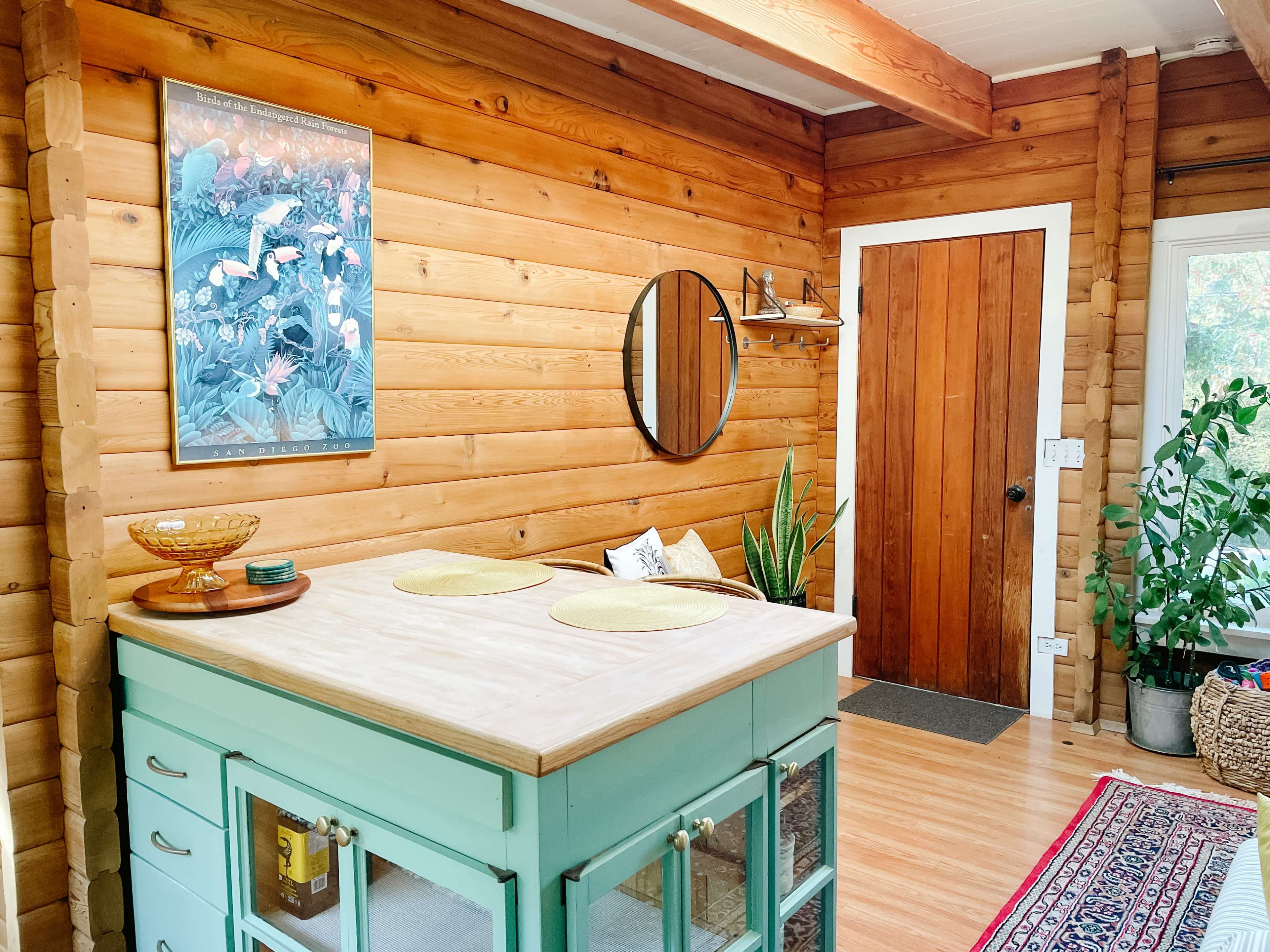 A cozy log cabin interior featuring a circular mirror, a wooden table with yellow placemats, and a doorway leading outside.