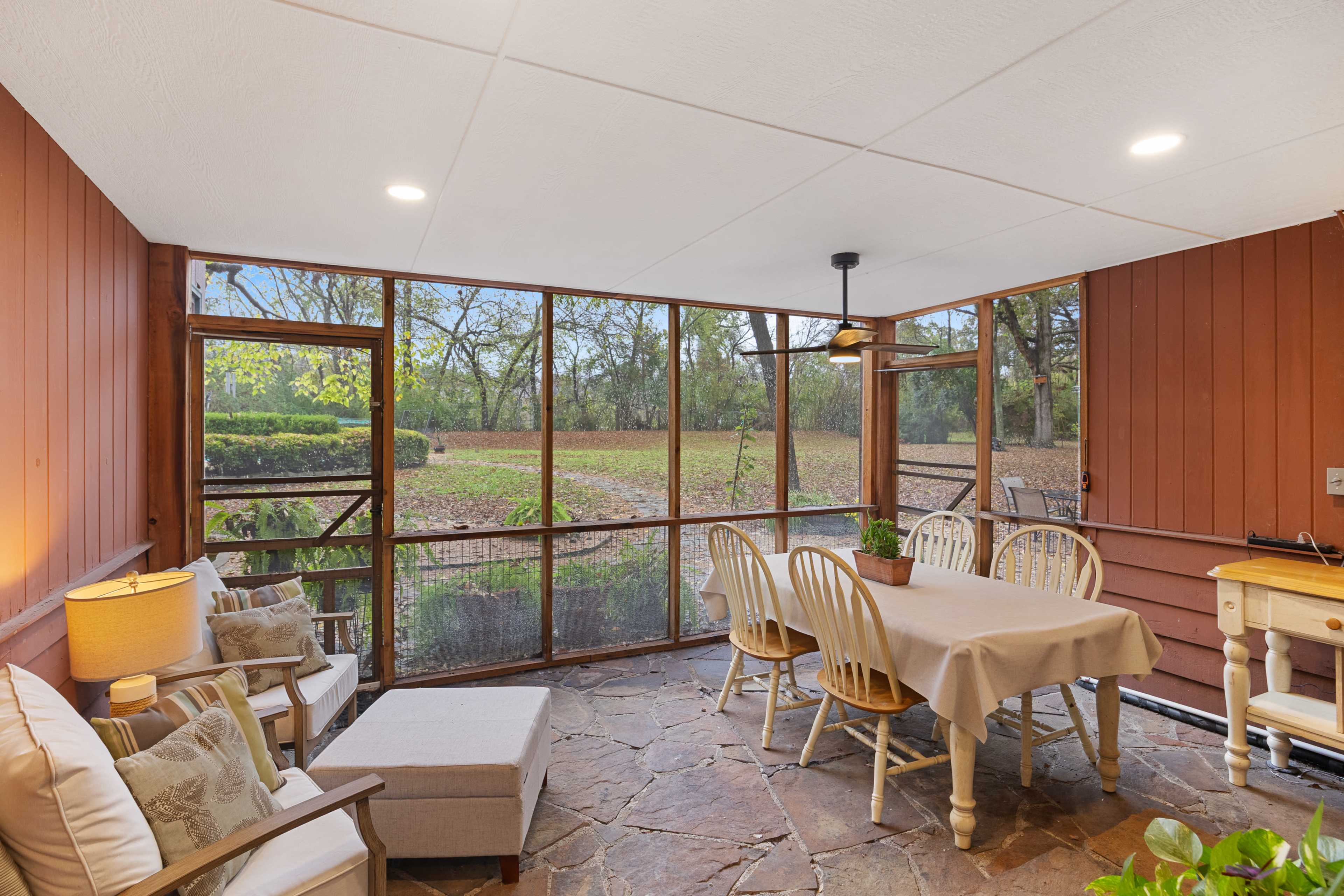 The image shows a screened-in porch featuring a dining table, seating area, and views of a wooded outdoor landscape.