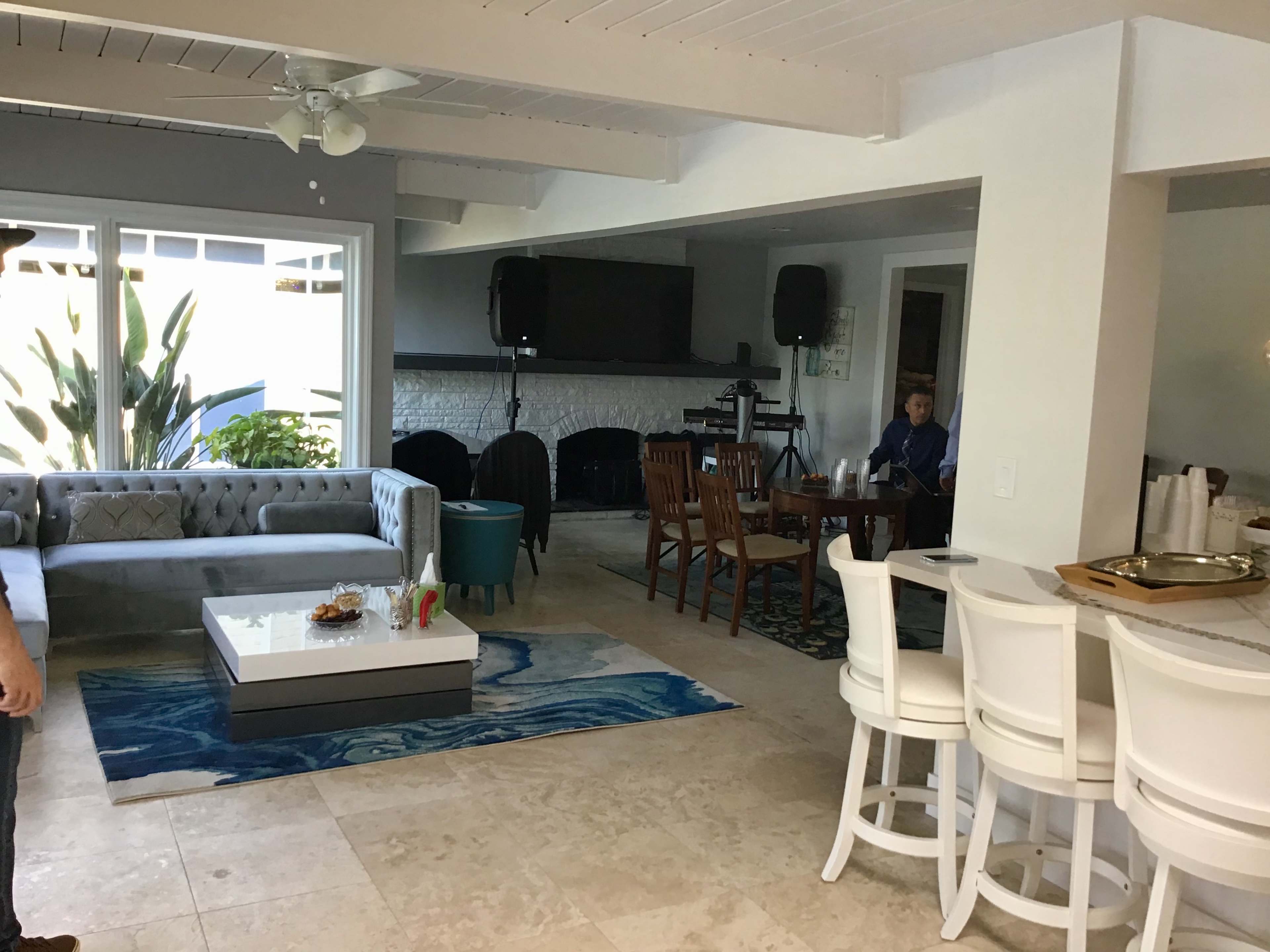 The image shows a spacious living area with a gray sofa, a coffee table, a dining area with wooden chairs, and a white bar with stools, surrounded by large windows and a tiled floor.