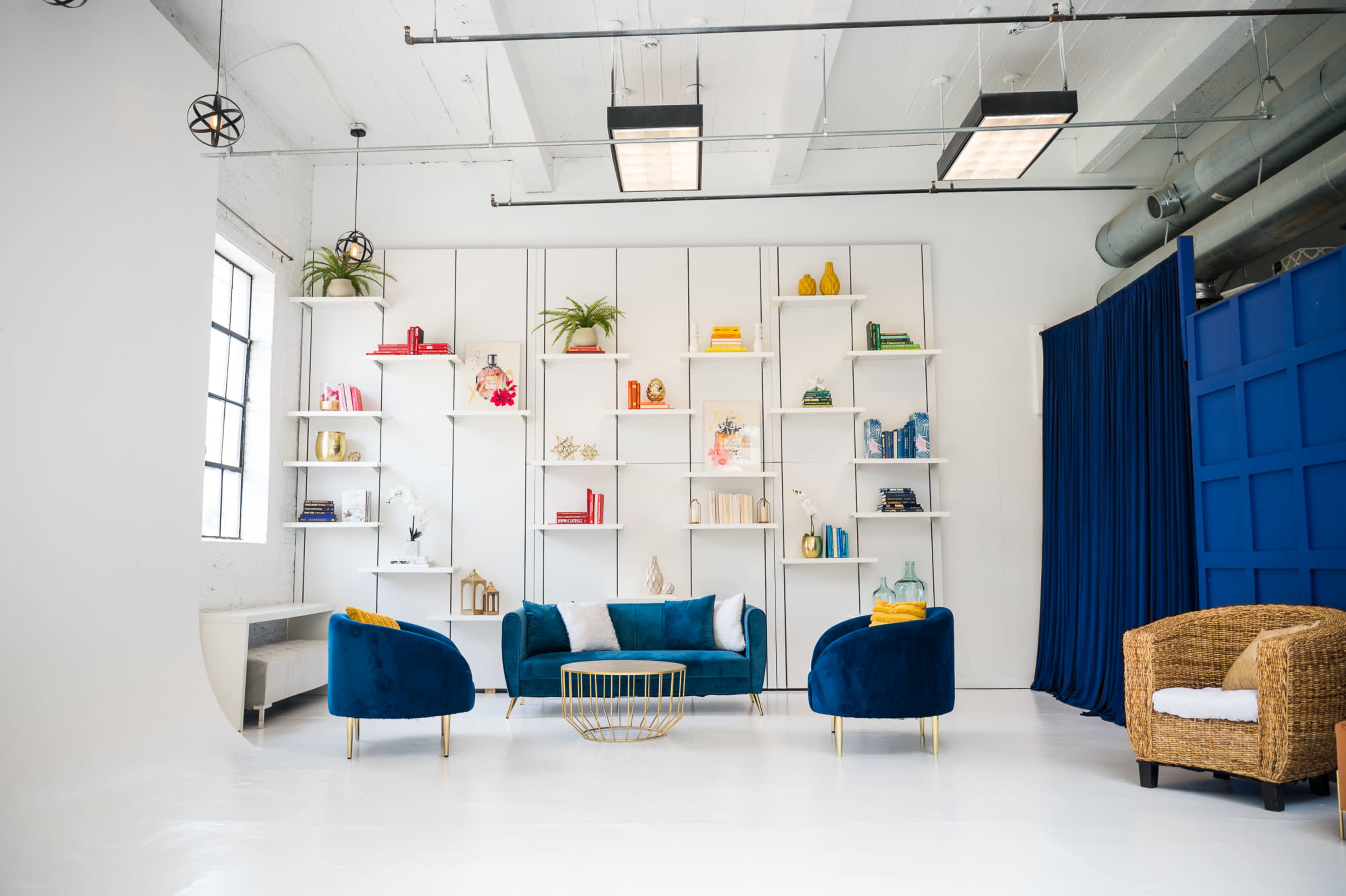 A modern interior showcases a bright room with a blue couch, two velvet chairs, a woven chair, and a white bookshelf filled with decorative items.