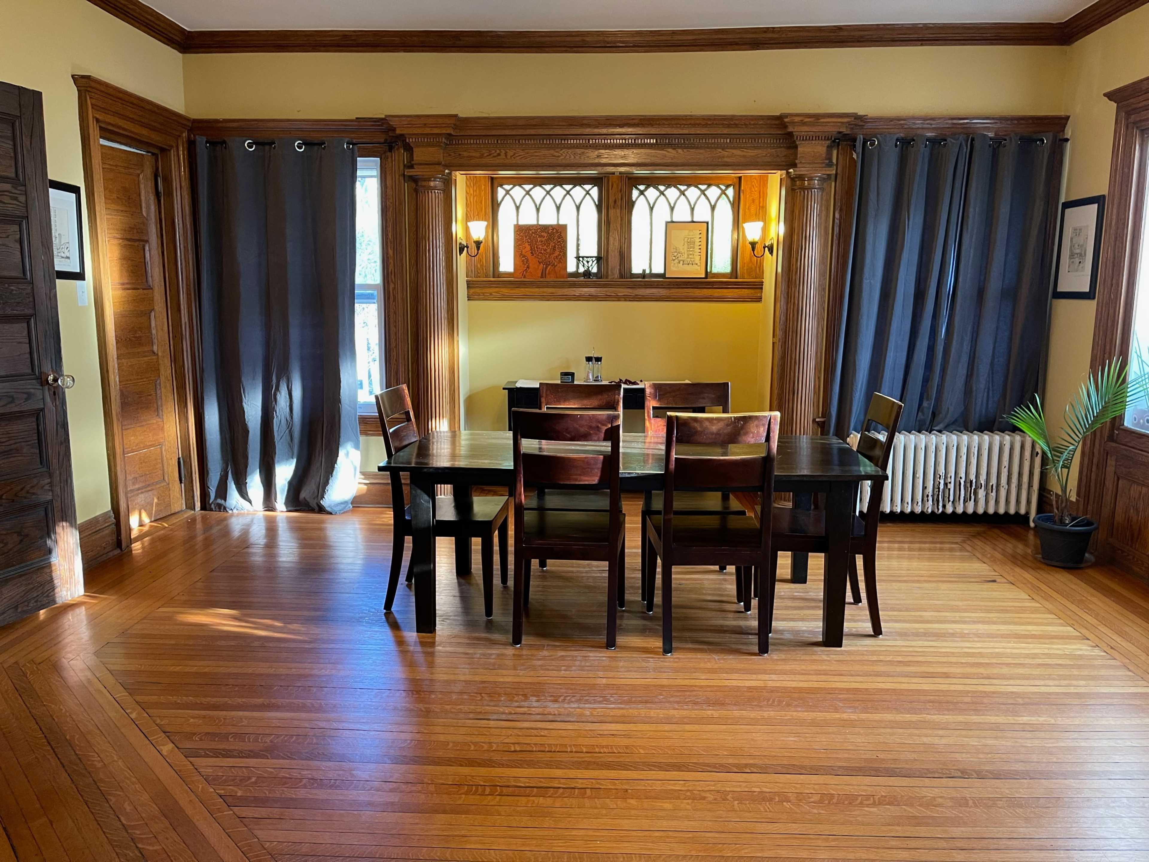 A wooden dining table surrounded by chairs is set in a sunlit room with large windows and dark curtains.