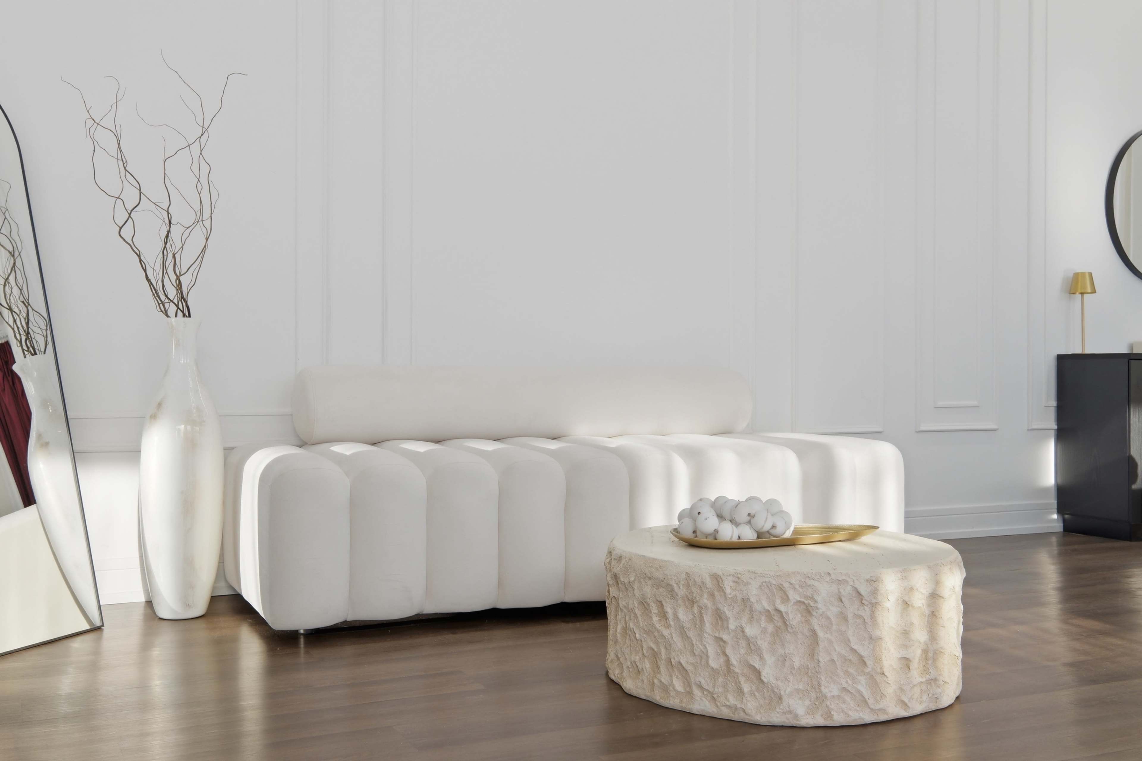 A minimalist living room features a white, curved sofa, a round coffee table with a textured surface, and decorative elements like a tall vase with branches.