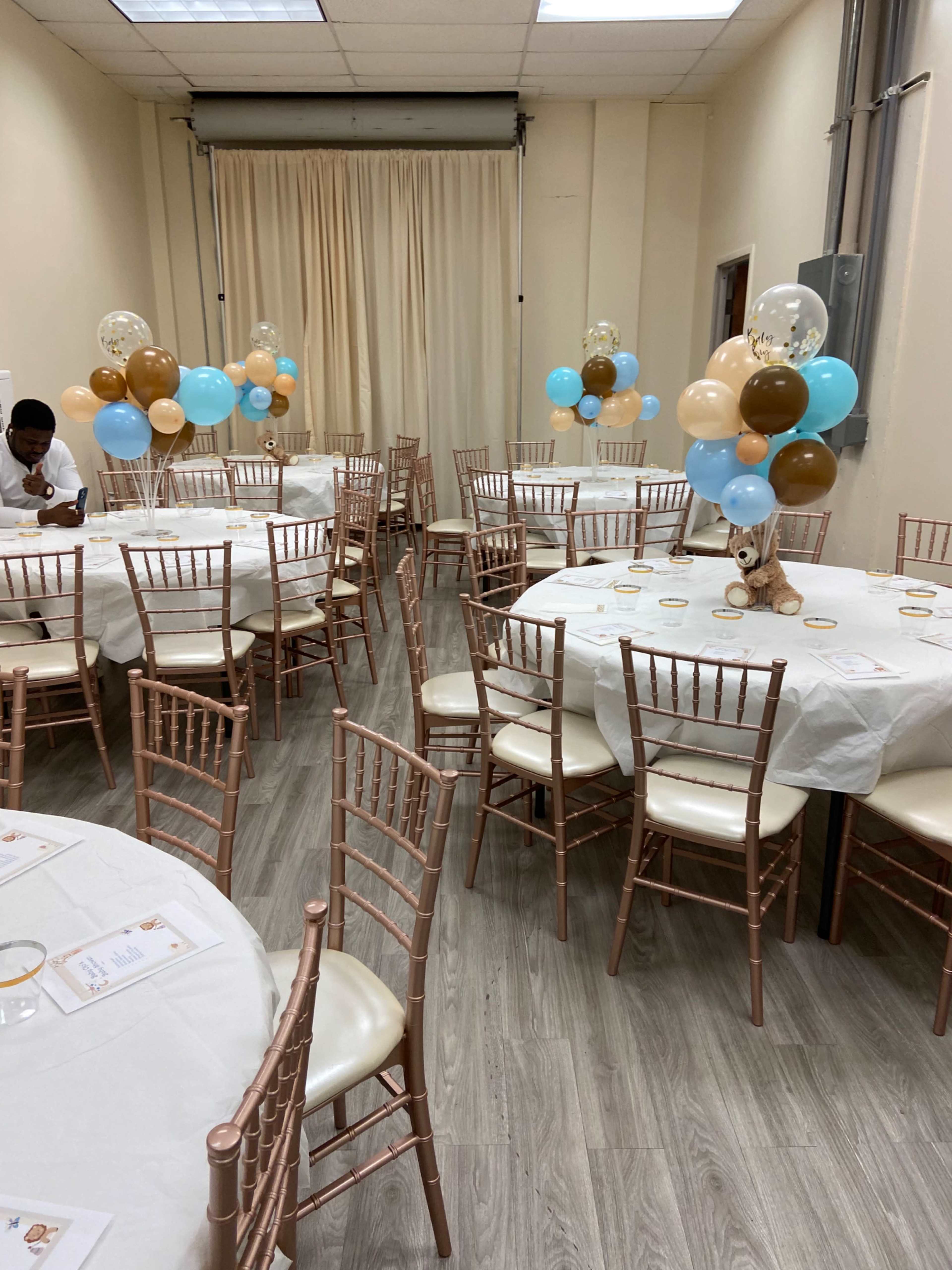 The room is set up for a celebration with white tables draped in tablecloths, surrounded by pastel balloons and rose gold chairs.
