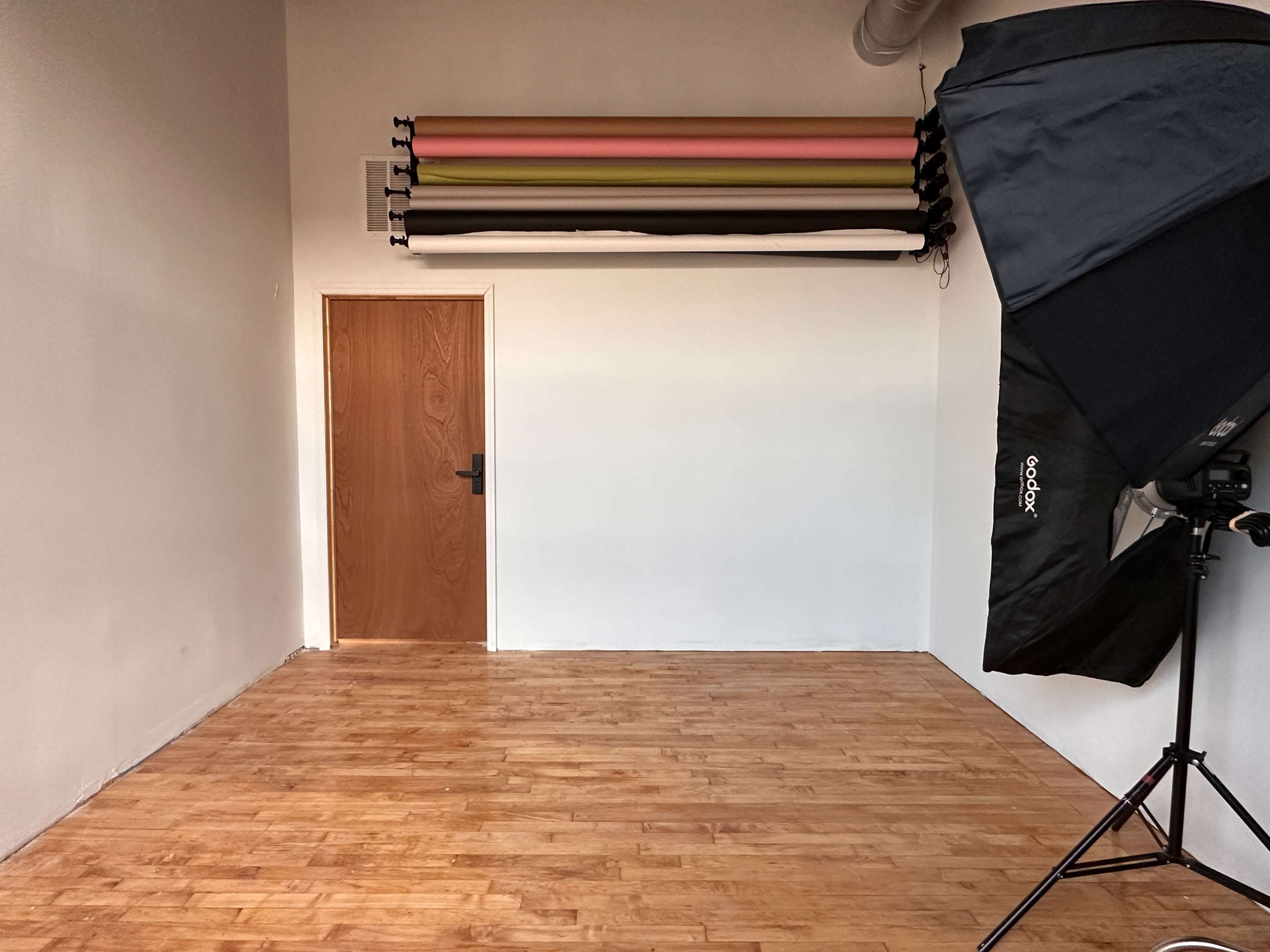A spacious photography studio with a wooden floor, a closed door, and a backdrop setup mounted on the wall.