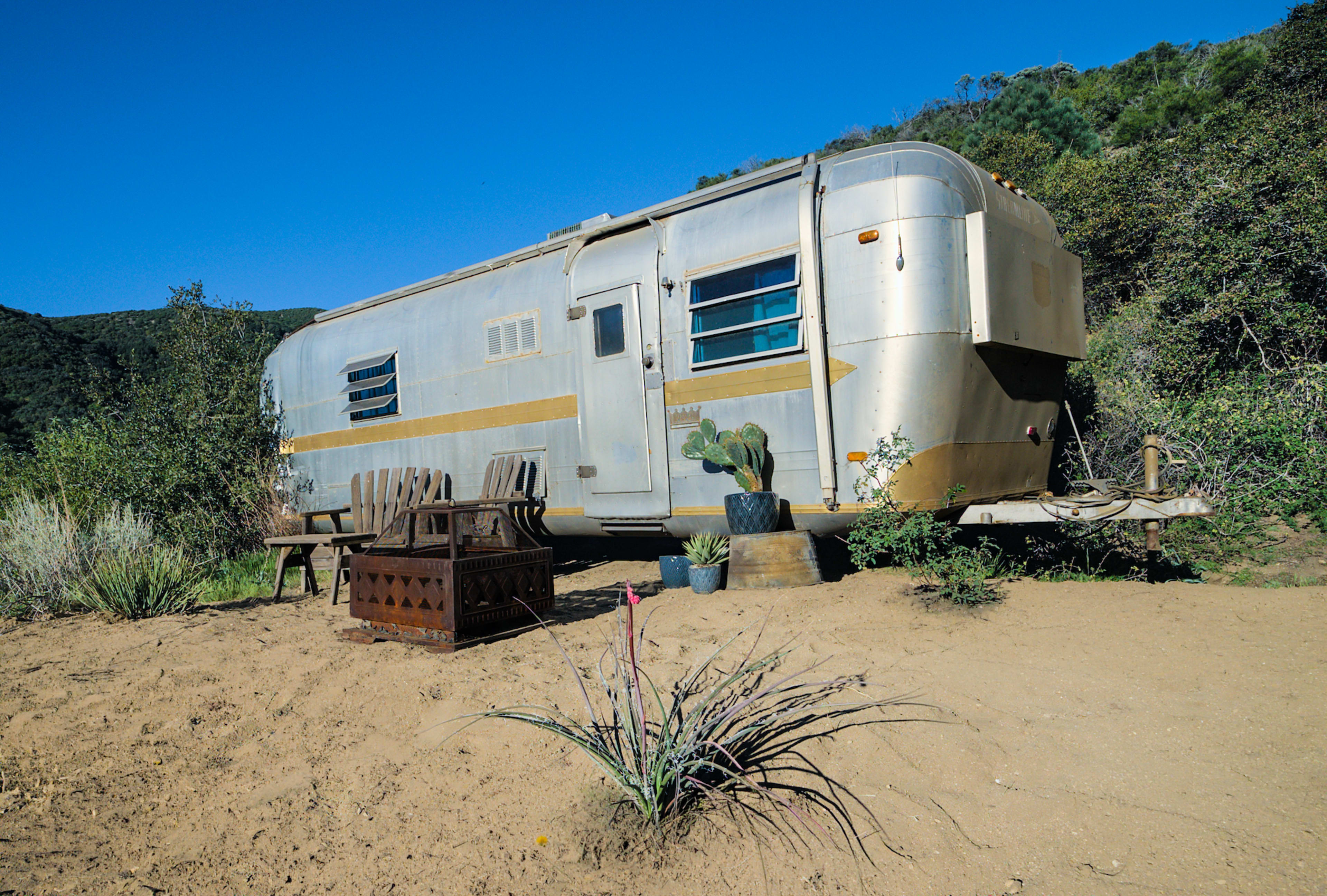 A vintage silver Airstream trailer is parked in a sandy area surrounded by green hills, with outdoor seating and potted plants nearby.