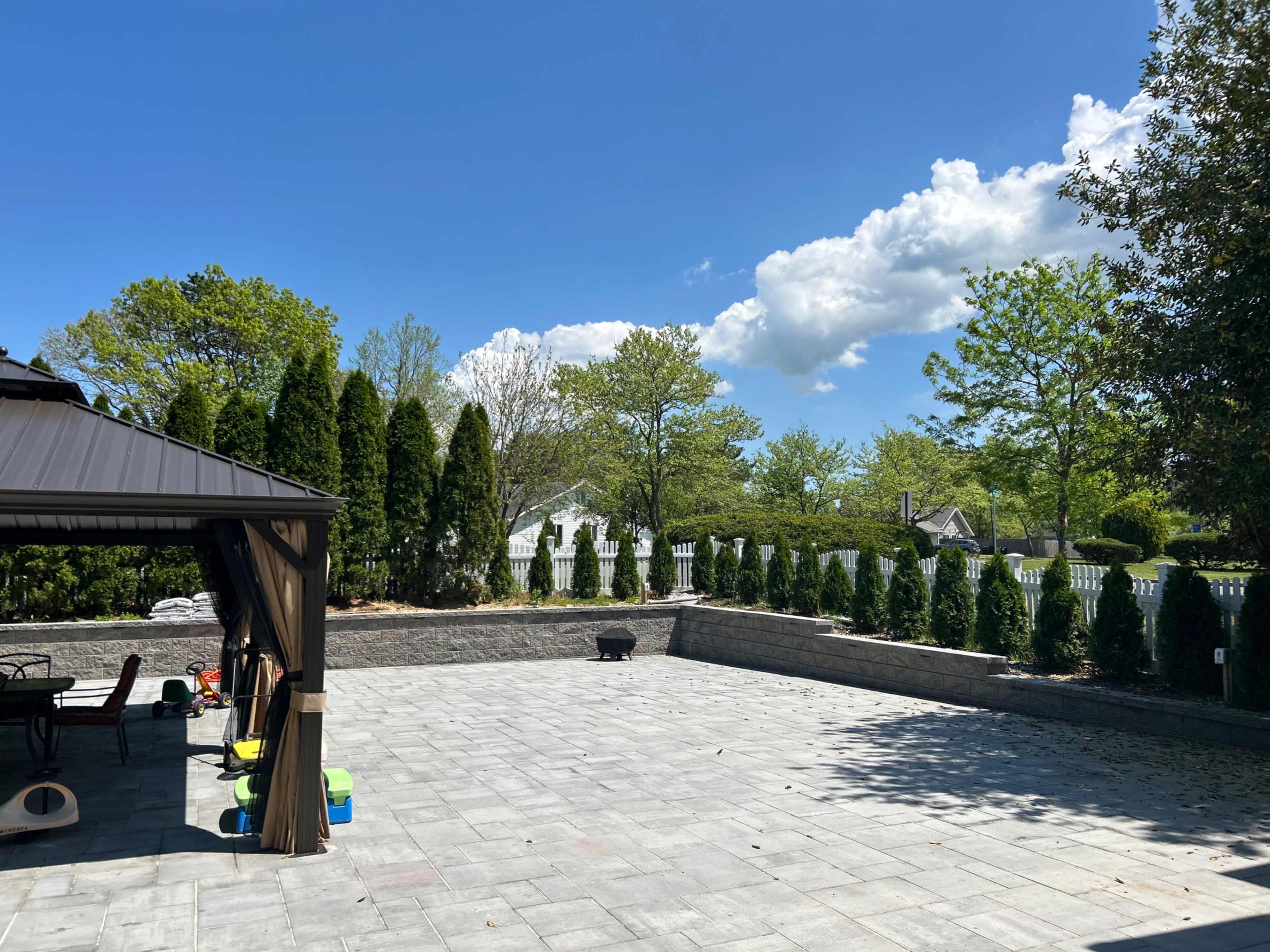 The image shows a spacious stone patio surrounded by manicured greenery and a clear blue sky with scattered clouds.