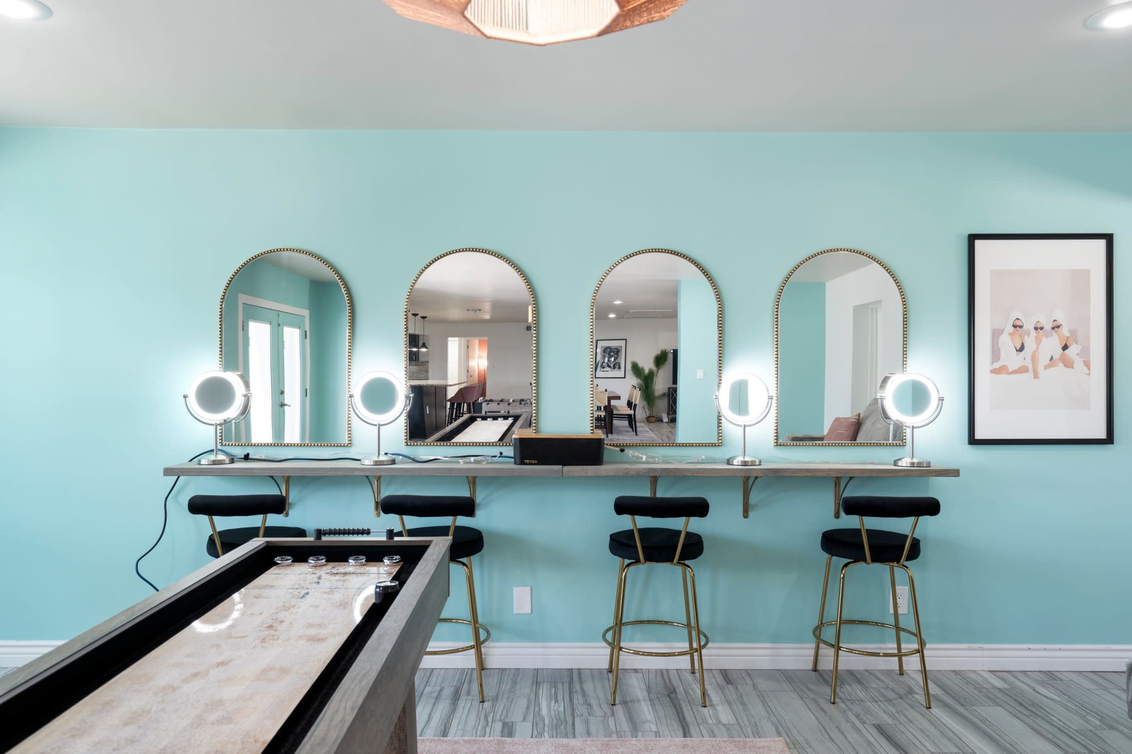 A bright room with a teal wall featuring a long table with three round mirrors and illuminated makeup stations, alongside a shuffleboard table.