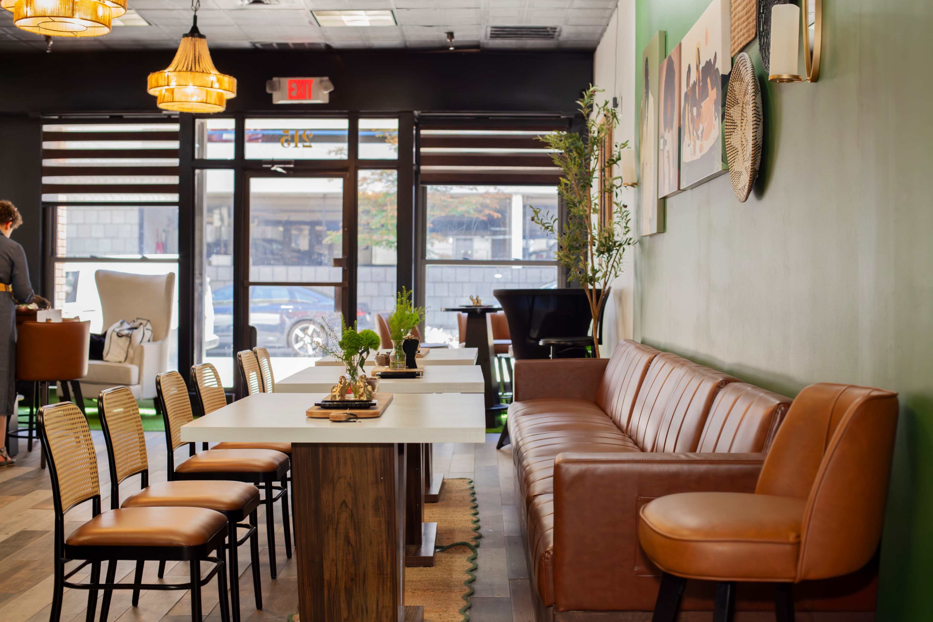 The image shows the interior of a café with a leather sofa, wooden tables, and stylish lighting, featuring a green accent wall and a view of the street through large windows.