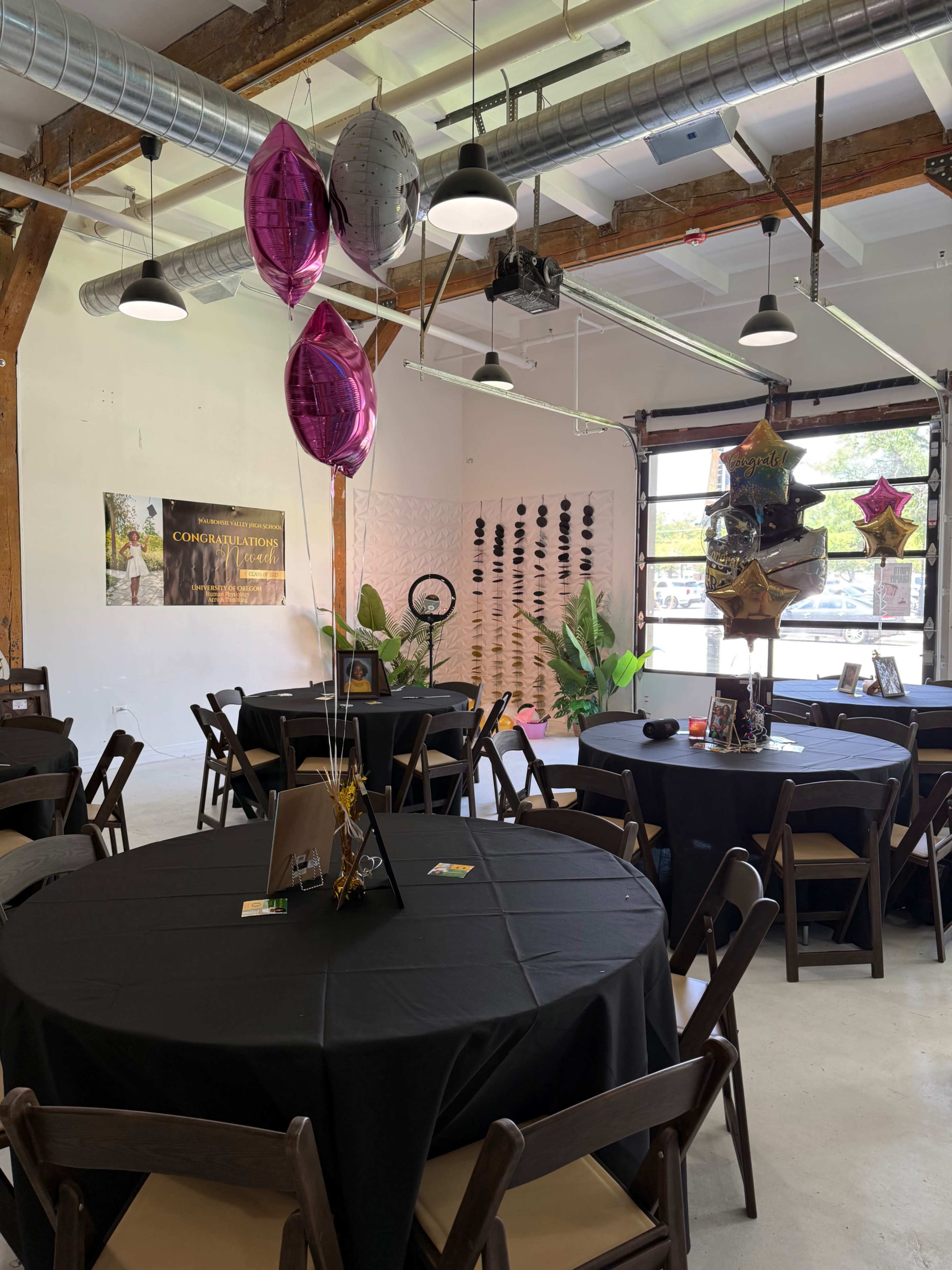 The image shows a decorated event space with round tables covered in black tablecloths, adorned with balloons and festive decorations.