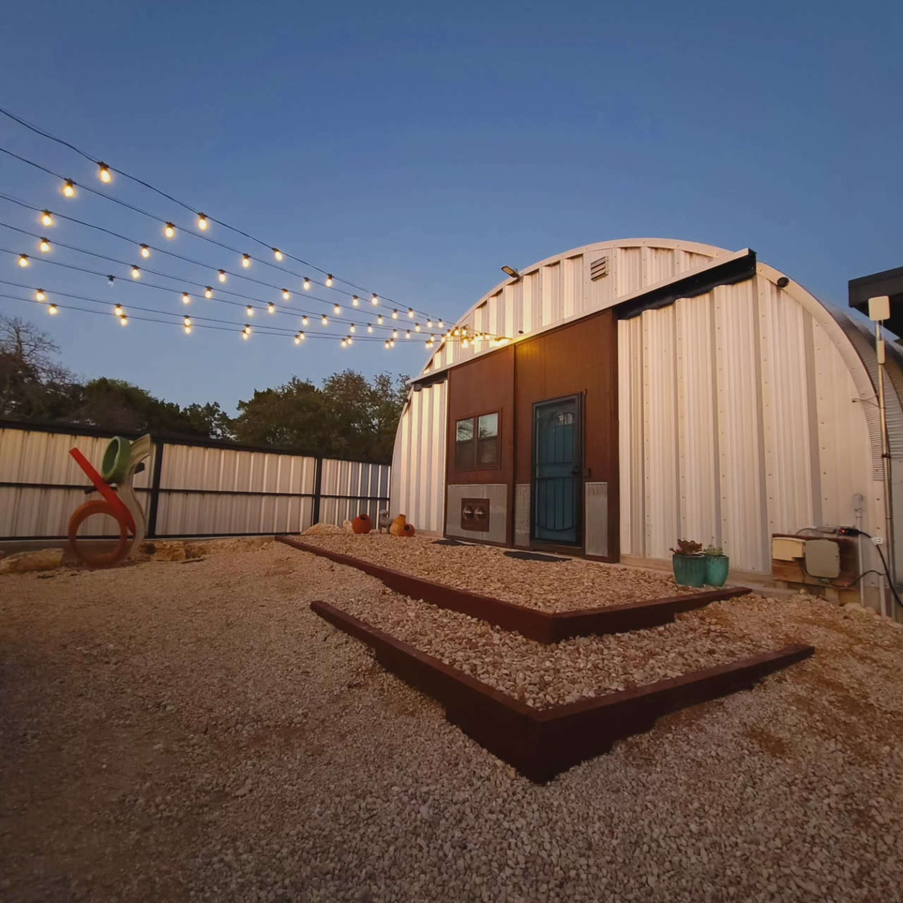 A round metal structure with a blue door is illuminated by string lights, surrounded by a gravel area and decorative elements.
