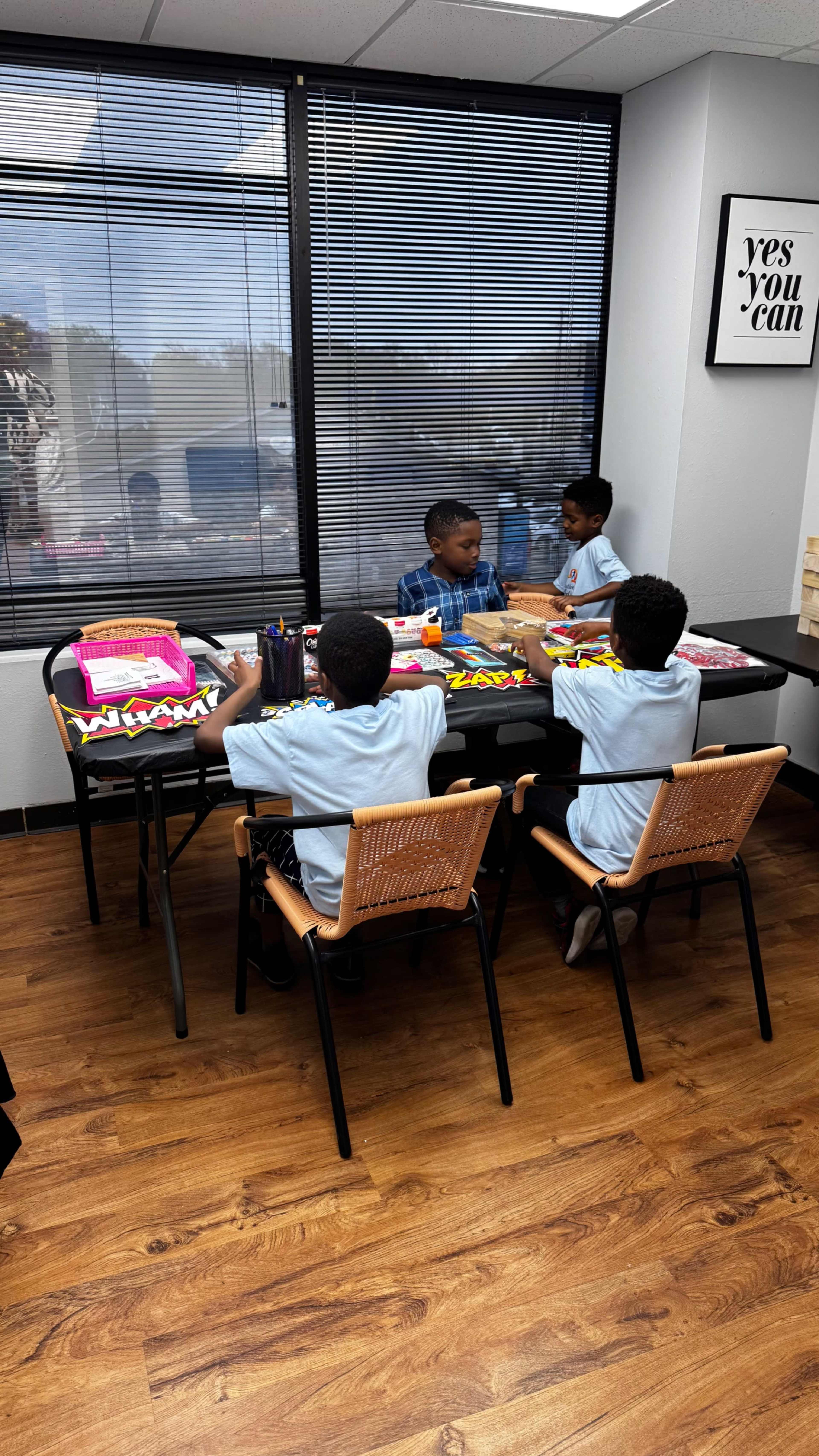 Three children are sitting at a table with art supplies, engaged in creative activities in a brightly lit room.