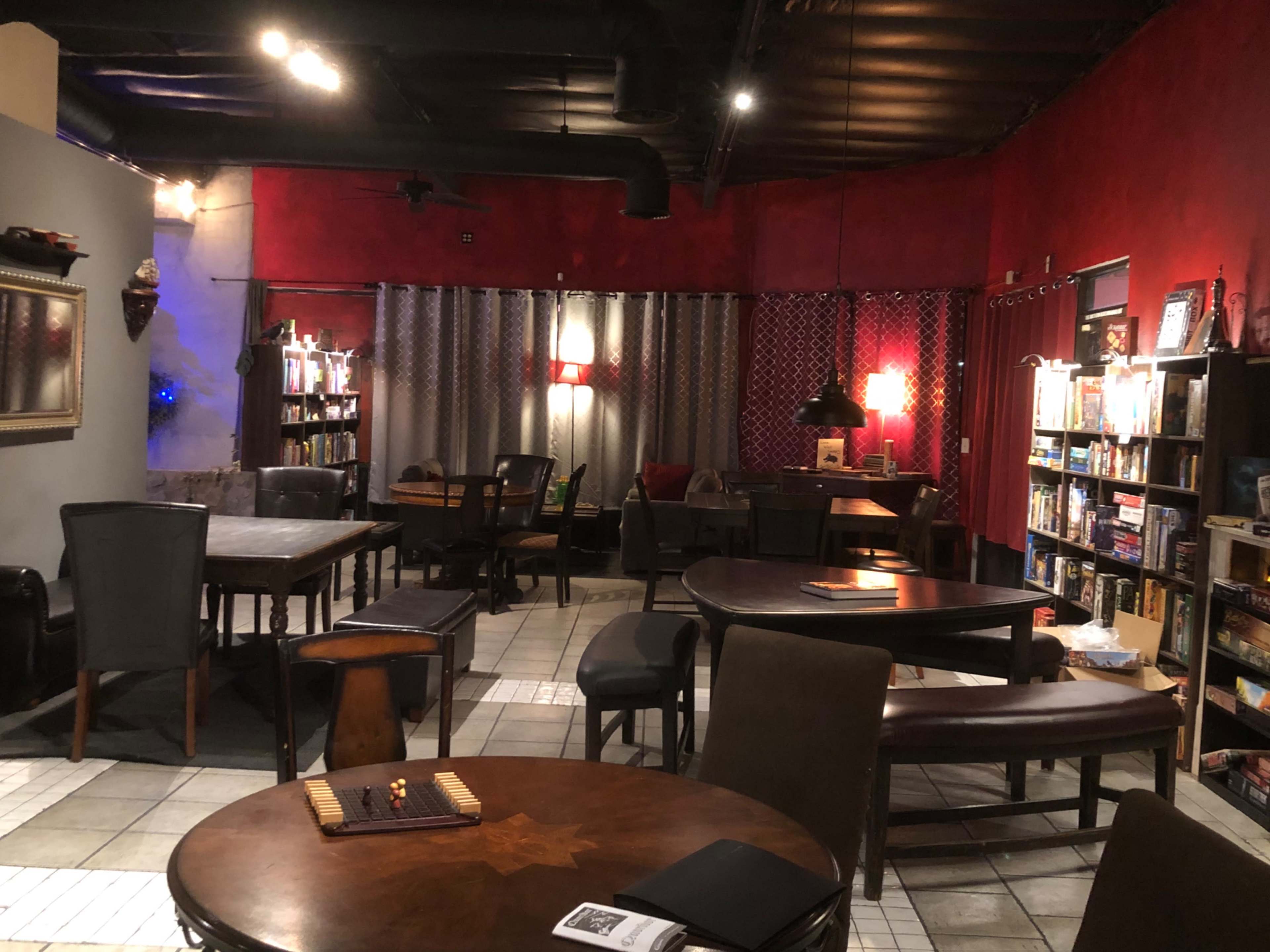 The image shows a cozy space with various tables and chairs, bookshelves filled with books, and red walls illuminated by warm lighting.