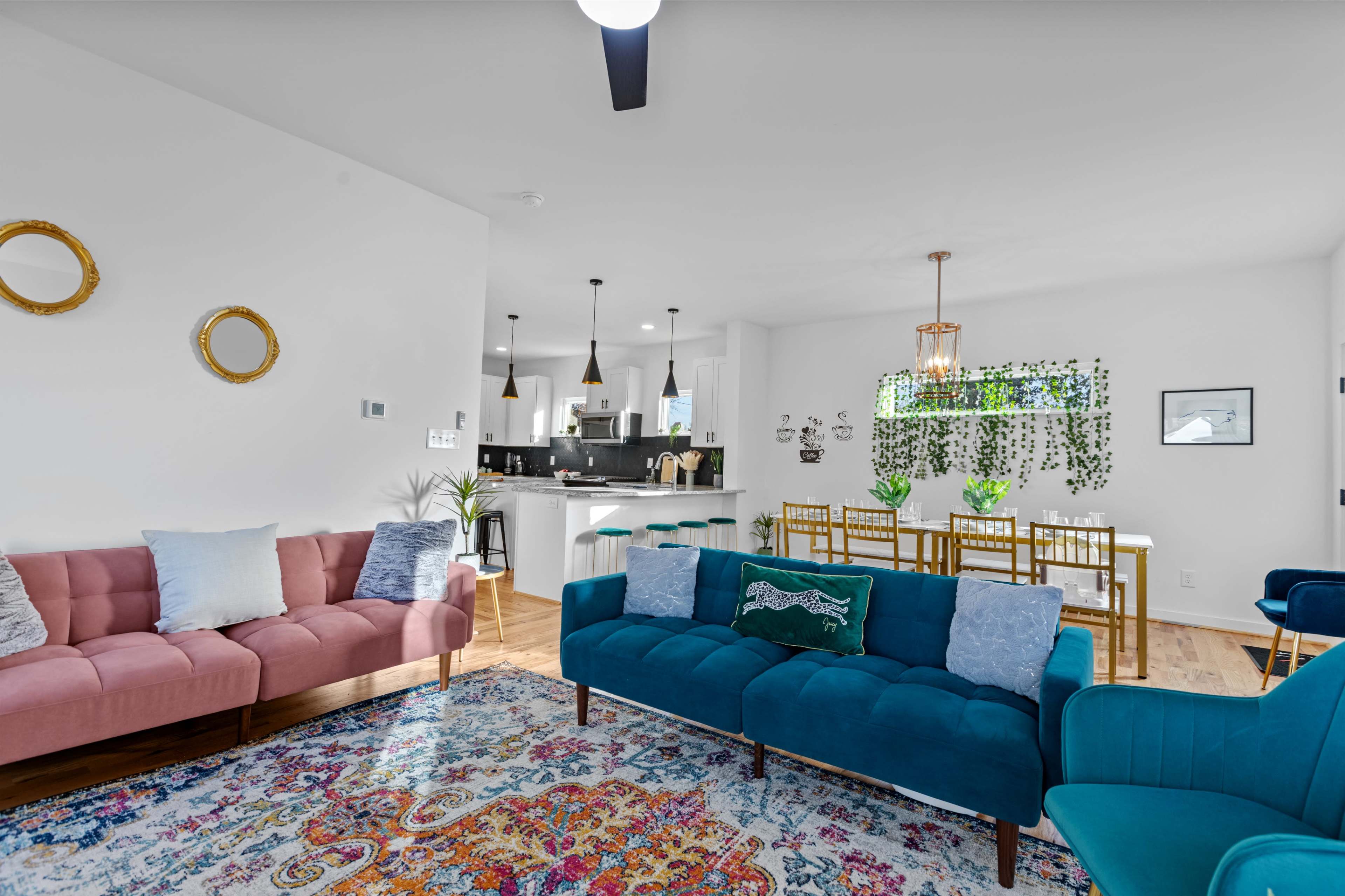 A modern living room with a mix of pink and blue sofas, a colorful area rug, and a dining area featuring a table with green decorative plants.