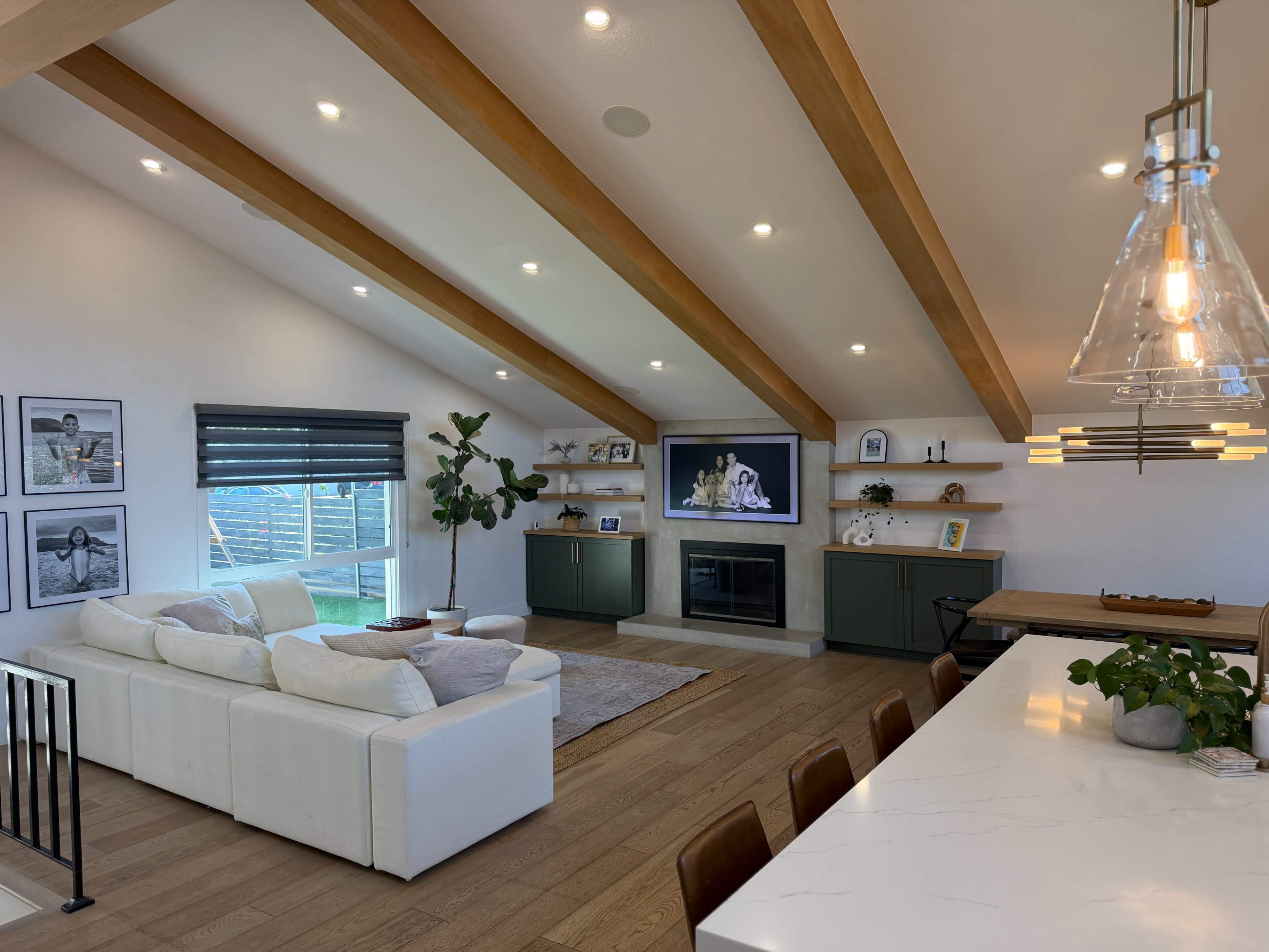 The image shows a modern living area with a white sectional sofa, a fireplace, green cabinetry, and wooden beams on the ceiling, complemented by a dining area with a marble countertop.