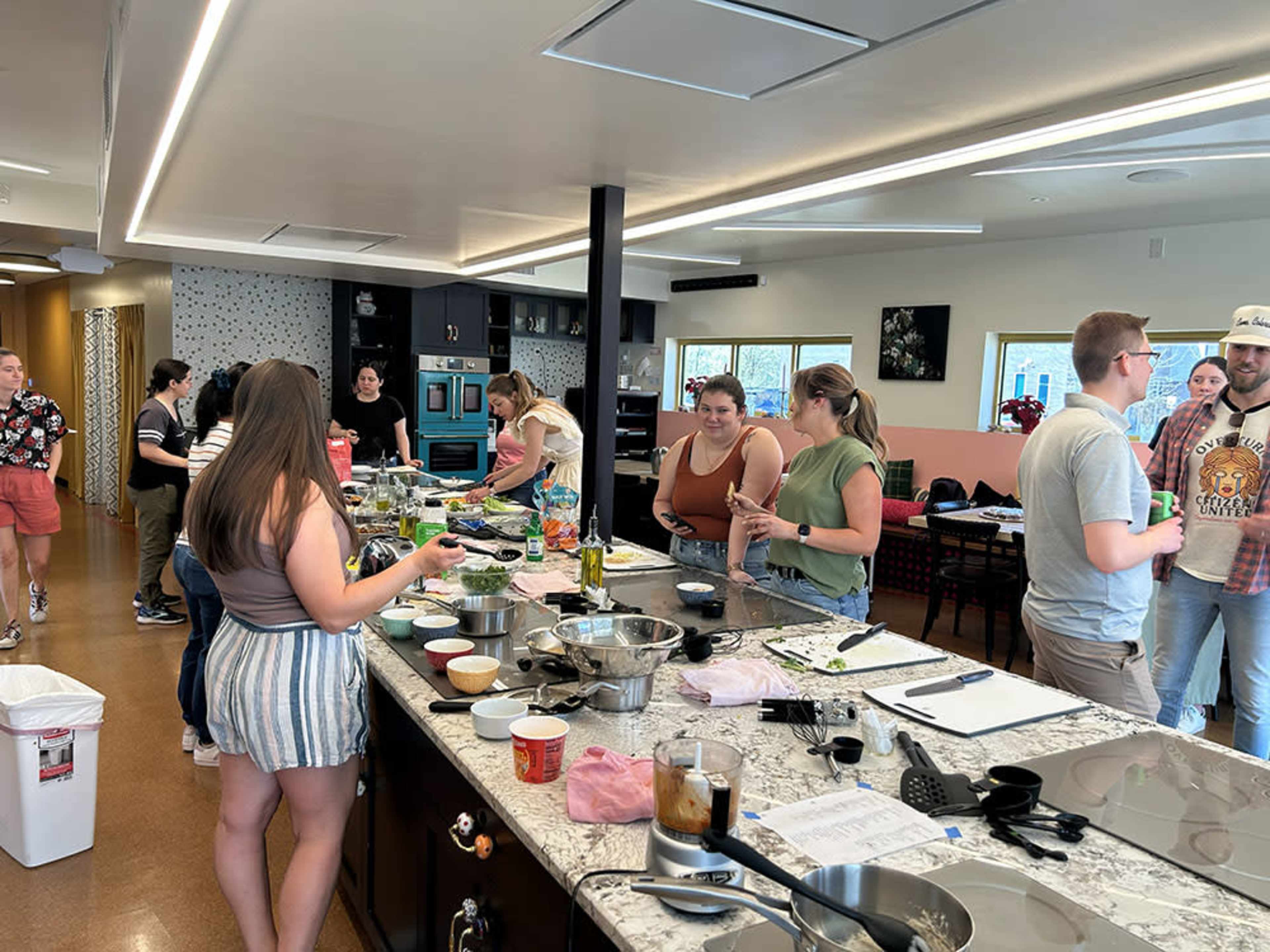 A group of people are cooking and preparing food in a large kitchen with various ingredients and utensils spread across a central island.