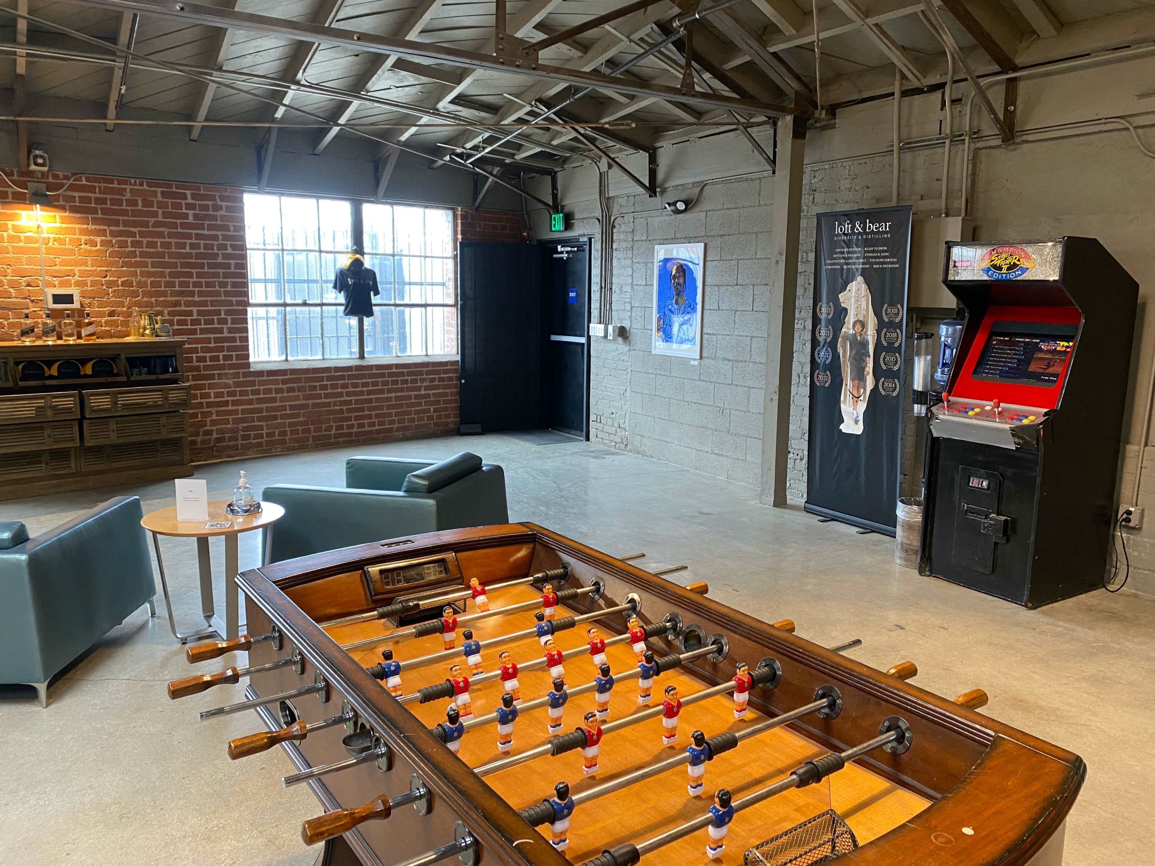 A game room with a foosball table at the center, seating areas, a vintage arcade game, and exposed brick walls.