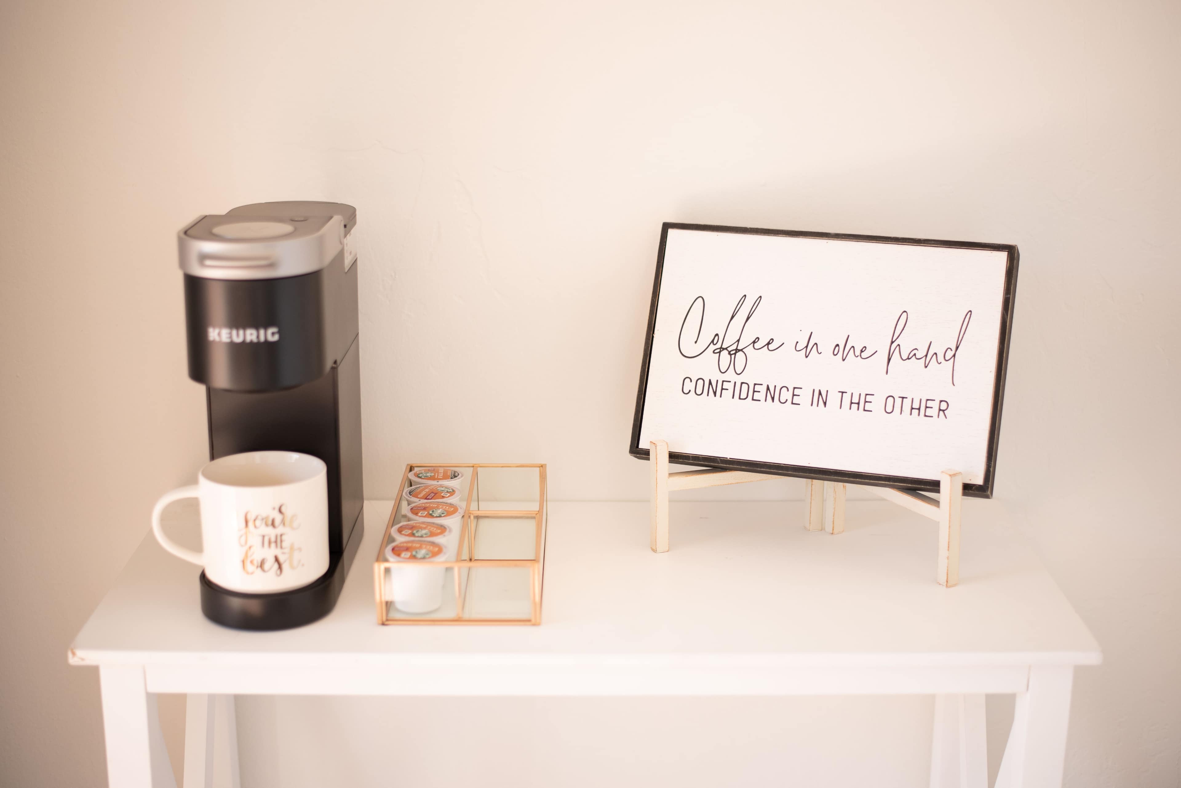 A coffee maker, a decorative sign, and a mug are displayed on a small table.