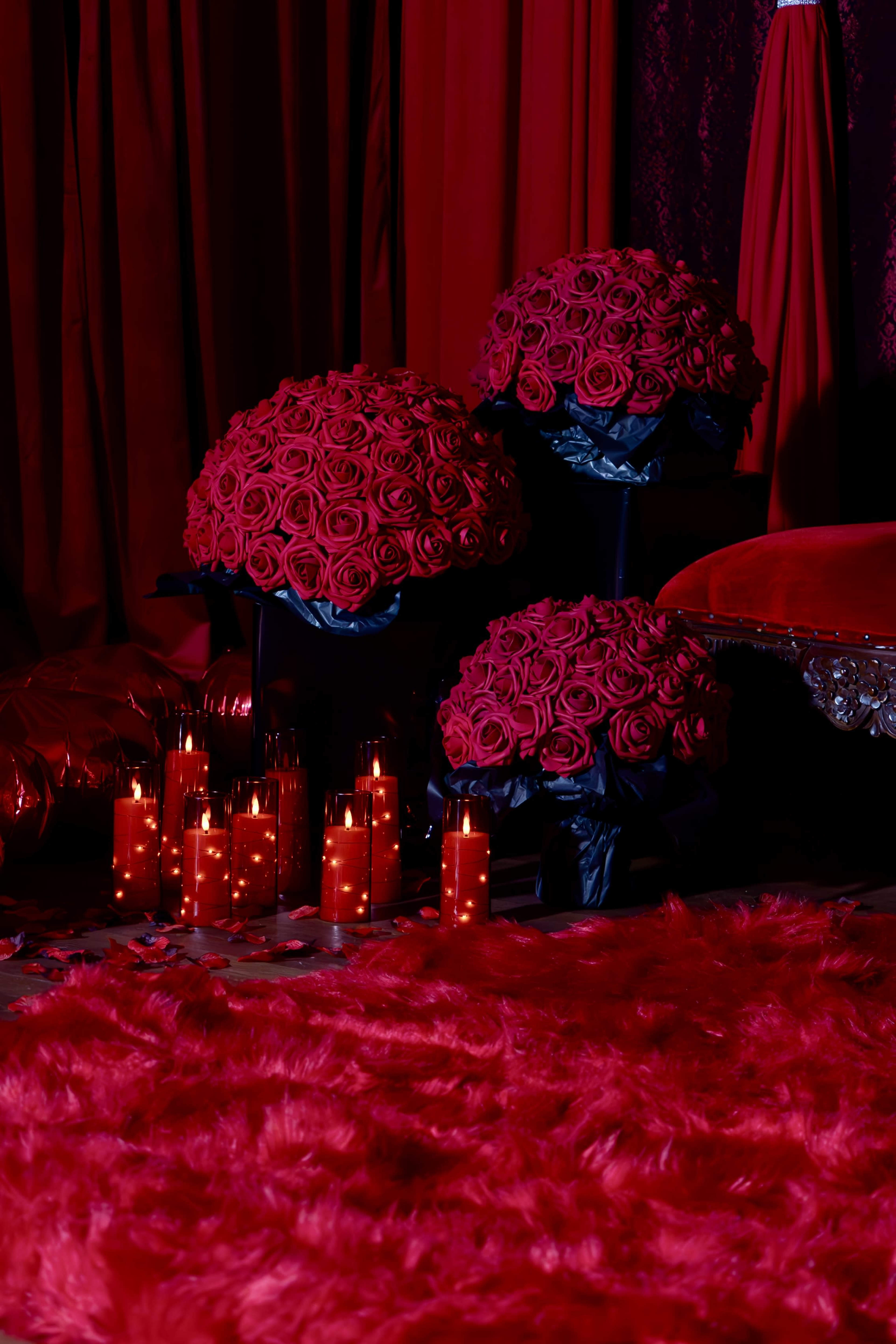Three large arrangements of deep red roses on black stands, surrounded by glowing candles, all set against a backdrop of red fabric and plush flooring.
