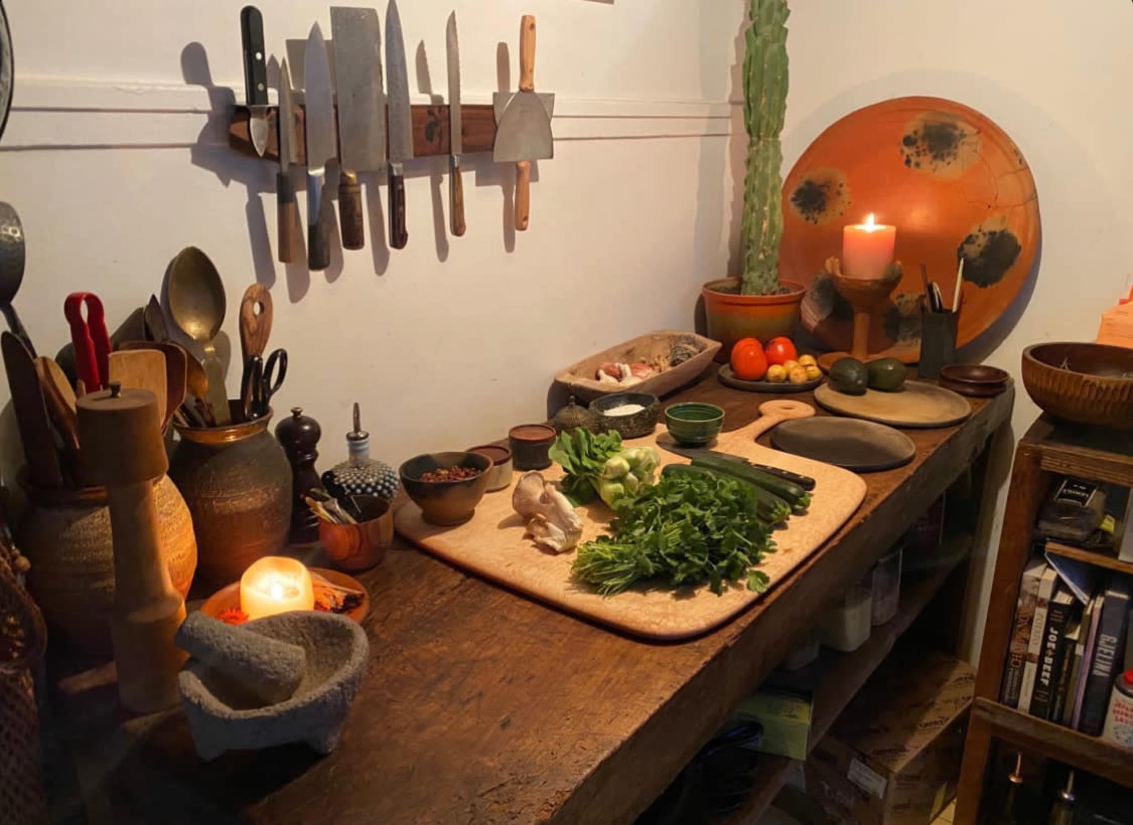 A wooden kitchen table is arranged with various cooking utensils, fresh vegetables, fruits, and candles, creating a rustic preparation space.
