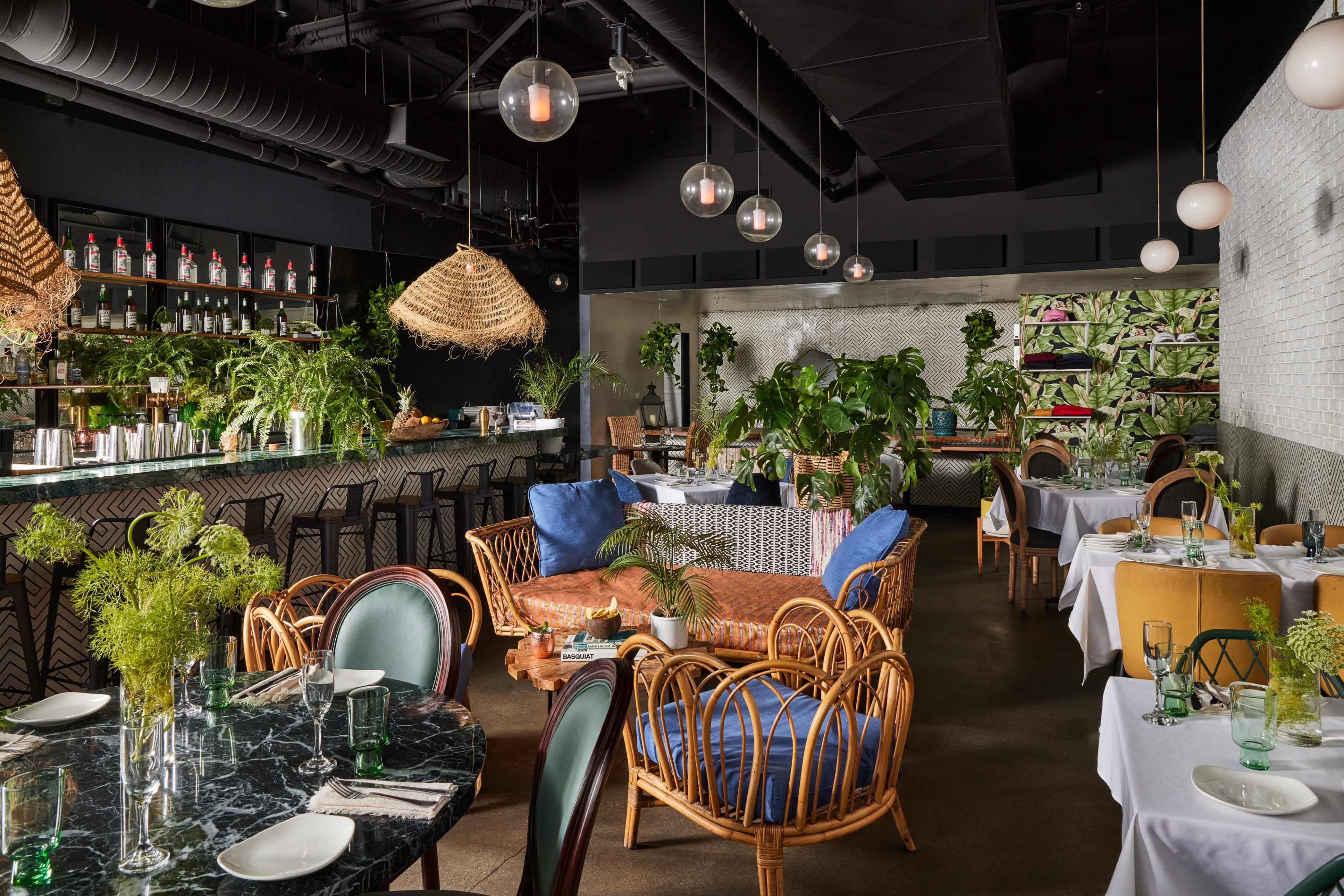The image depicts a modern restaurant interior with a bar area, seating arrangements featuring rattan furniture, decorative plants, and pendant lights hanging from the ceiling.