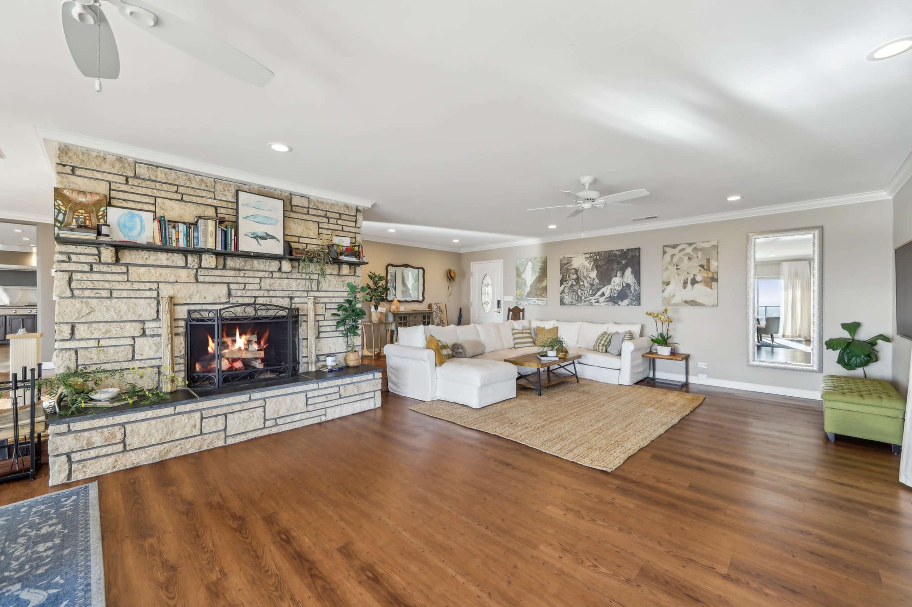 The living room features a large stone fireplace, a white sectional sofa, and a natural fiber rug, with art pieces on the walls and plants throughout.