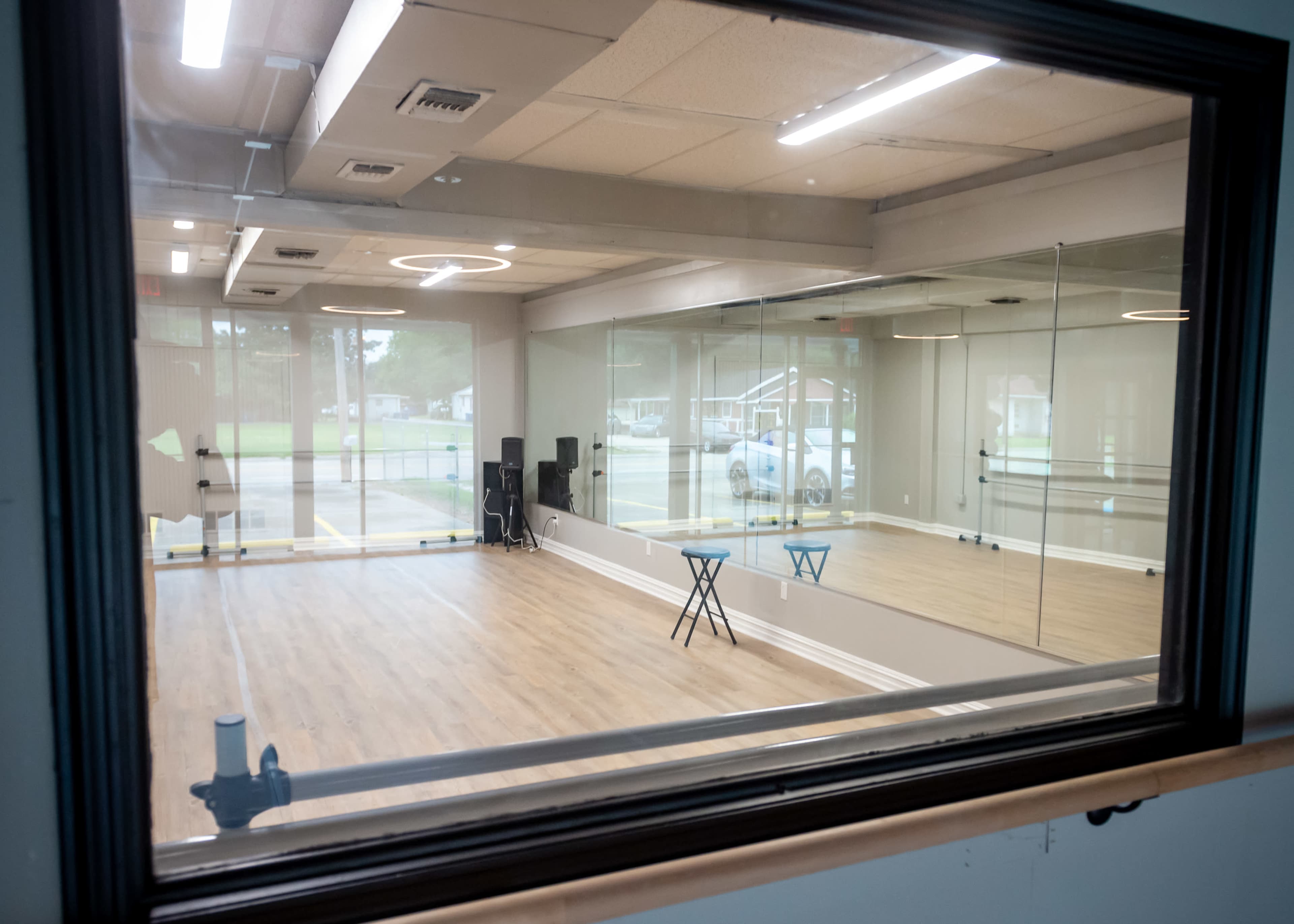The image shows a bright, empty dance studio with wooden flooring and large mirrors, viewed through a window.