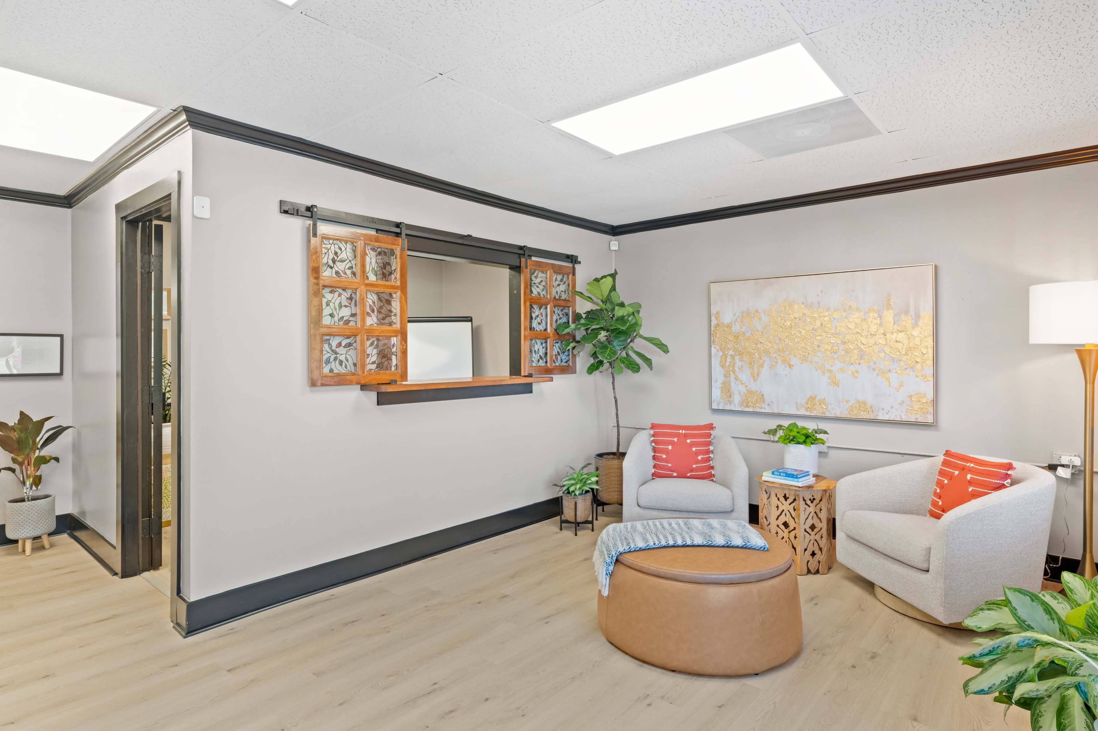 A modern waiting area with two chairs, a round ottoman, a plant, and a decorative wall art piece.