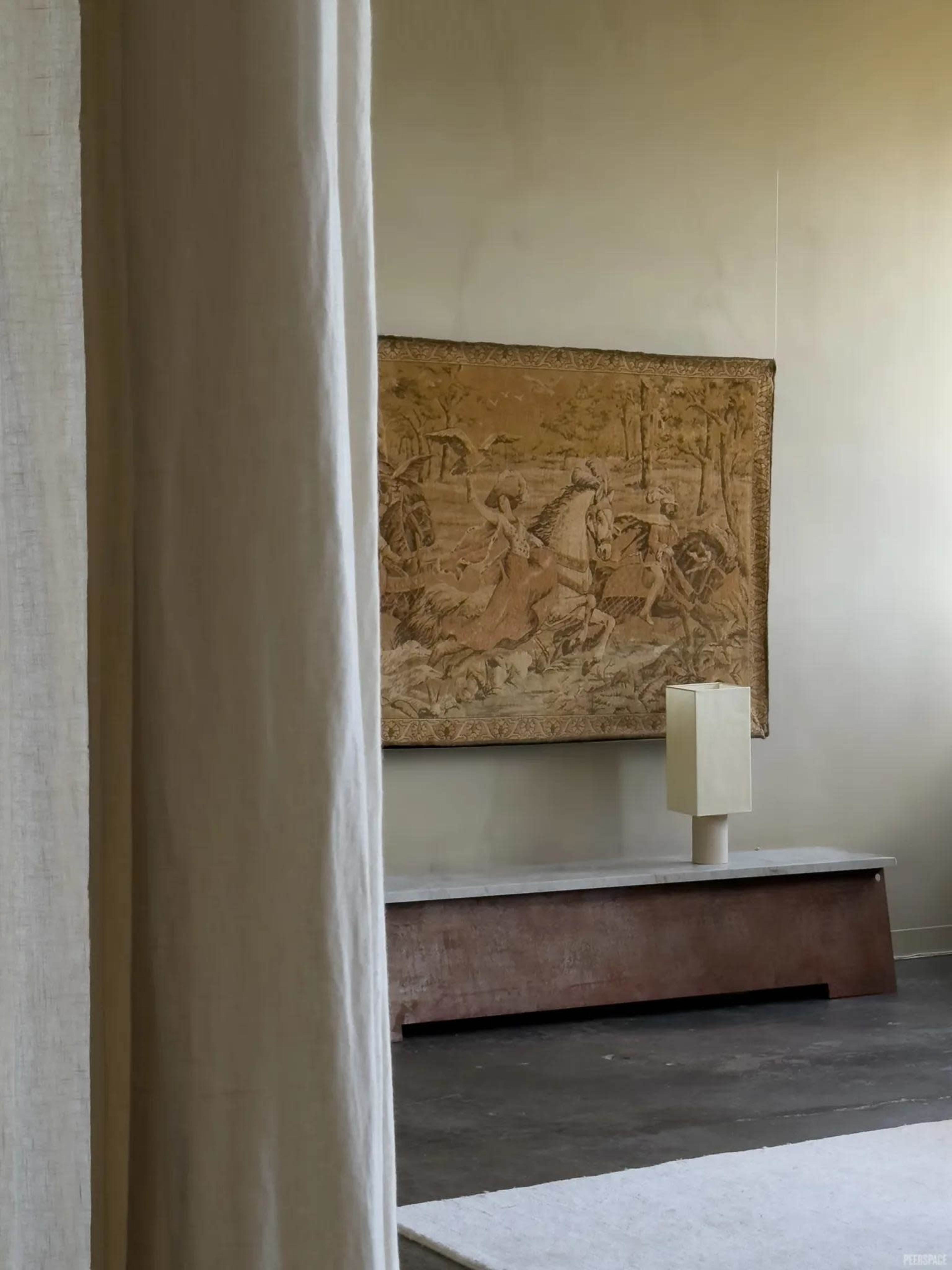 The image shows a minimalist interior featuring a tapestry on the wall, a low wooden bench, and a simple lamp in a softly lit room.