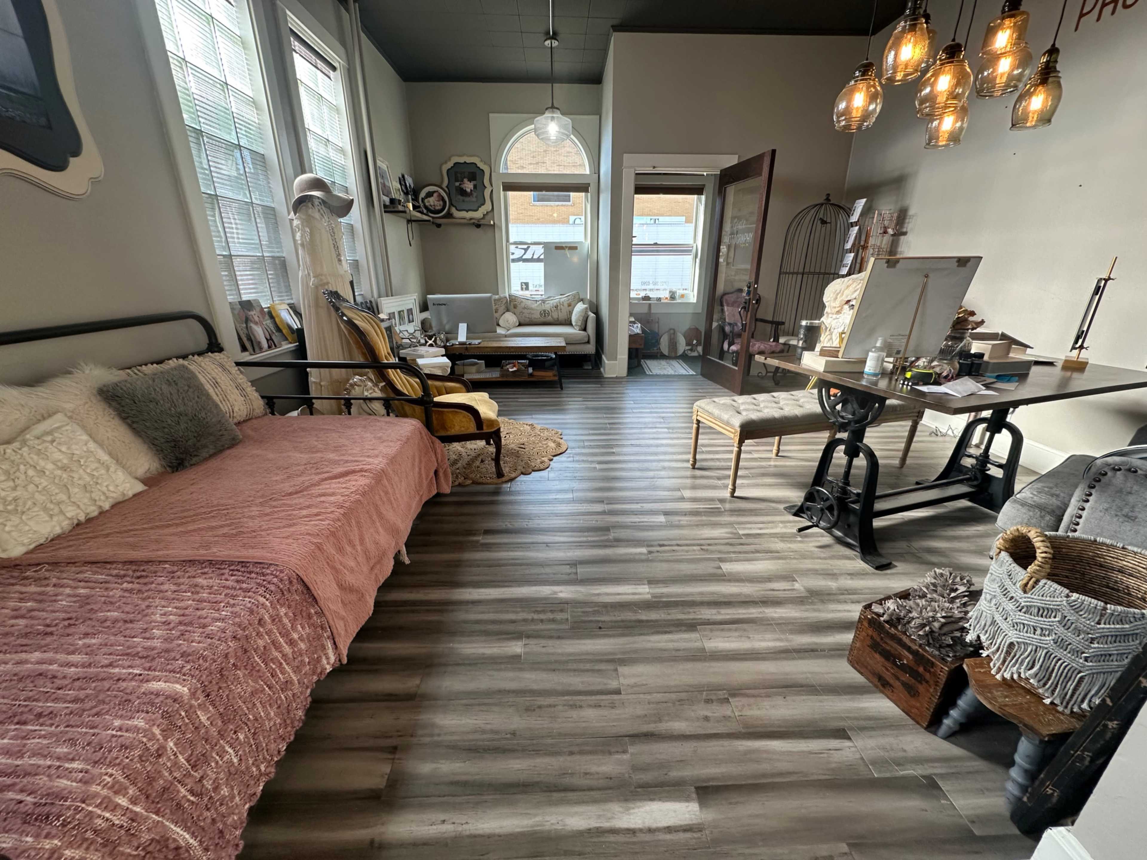 The image shows a cozy interior space featuring a combination of seating areas, a workspace, and decorative elements with large windows allowing natural light.