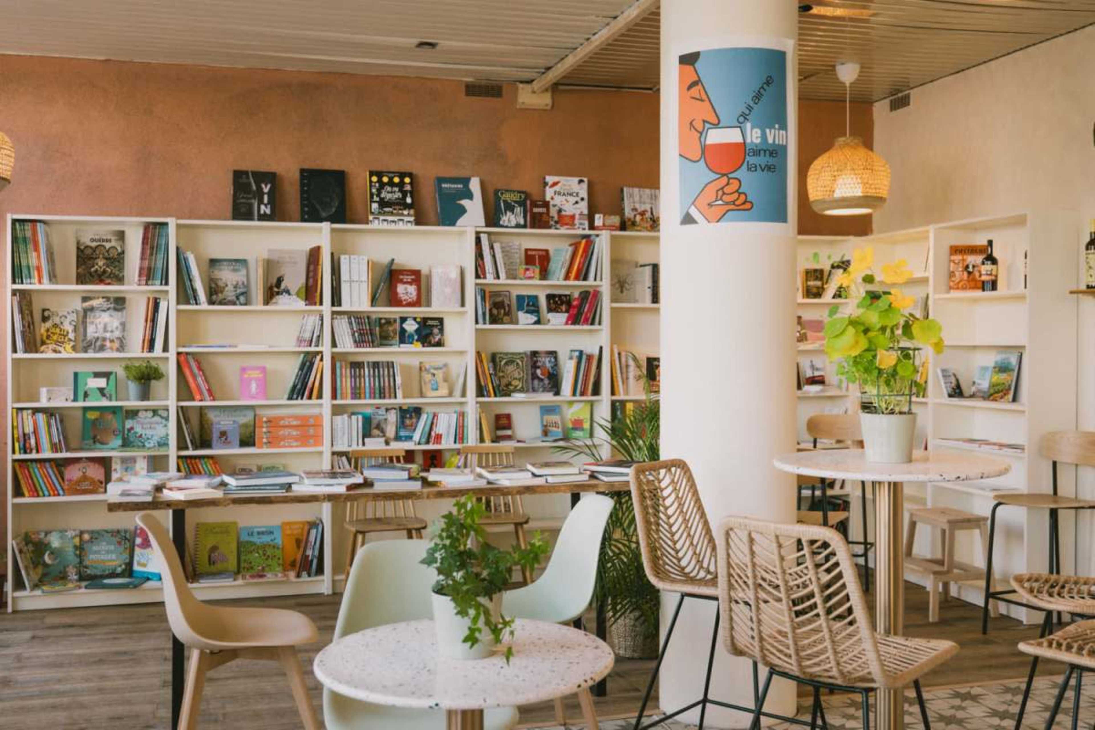 The image shows a bright, modern café with bookshelves filled with colorful books and small tables surrounded by wicker chairs.