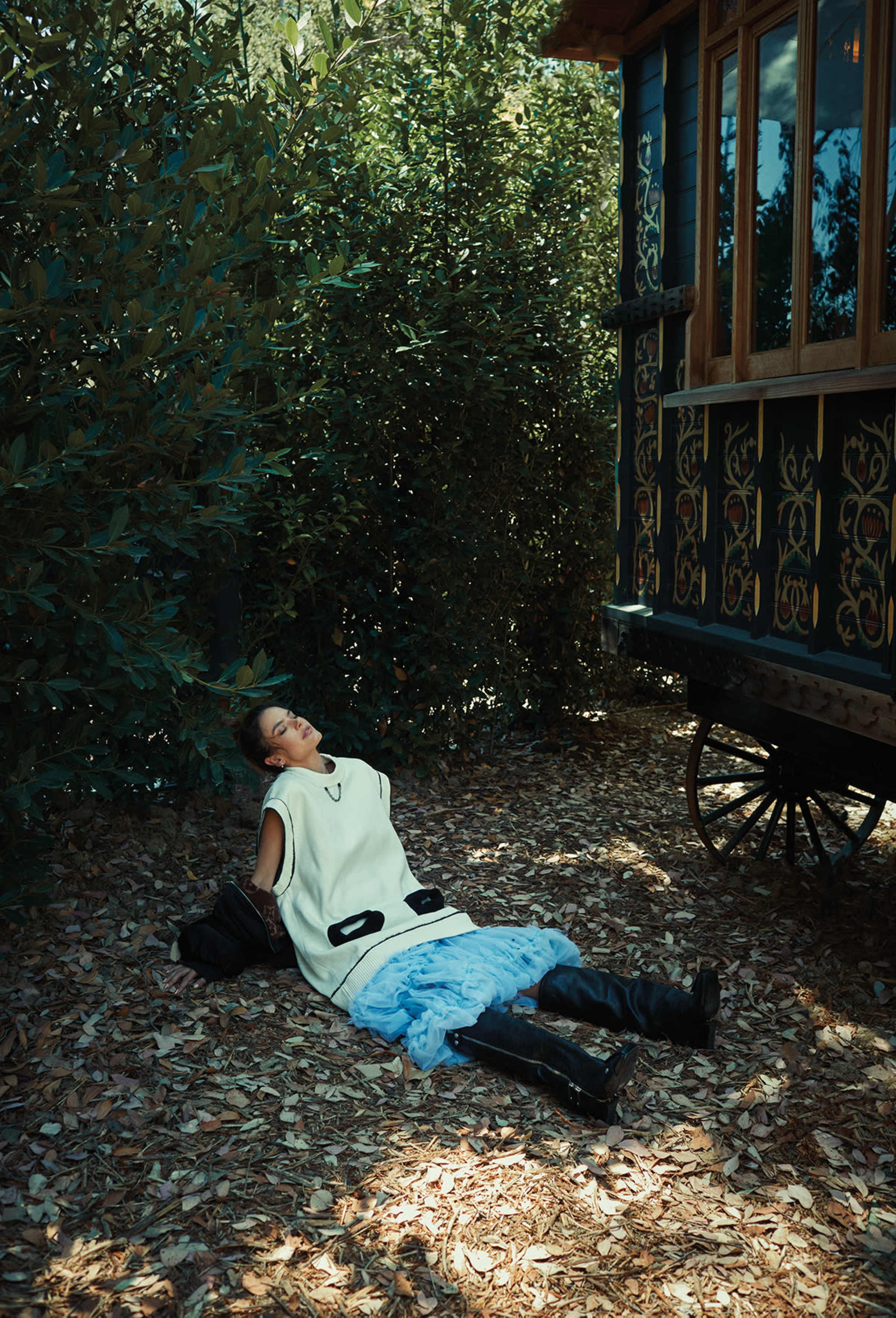 A person sits on the ground near a decorated trailer, surrounded by dense foliage and scattered leaves.