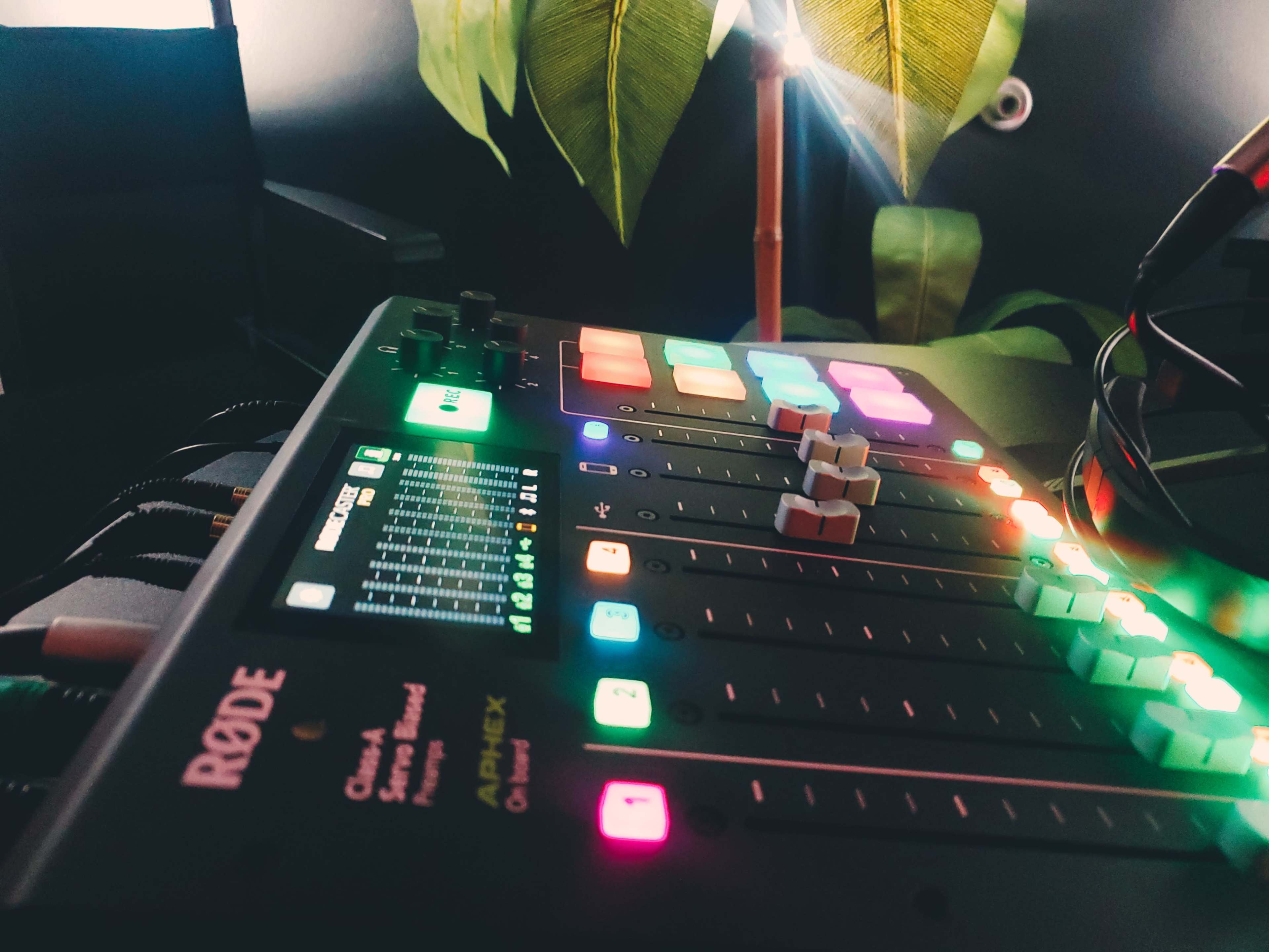 A Rode audio mixing console with colorful buttons and sliders, set against a backdrop of green leaves and soft lighting.