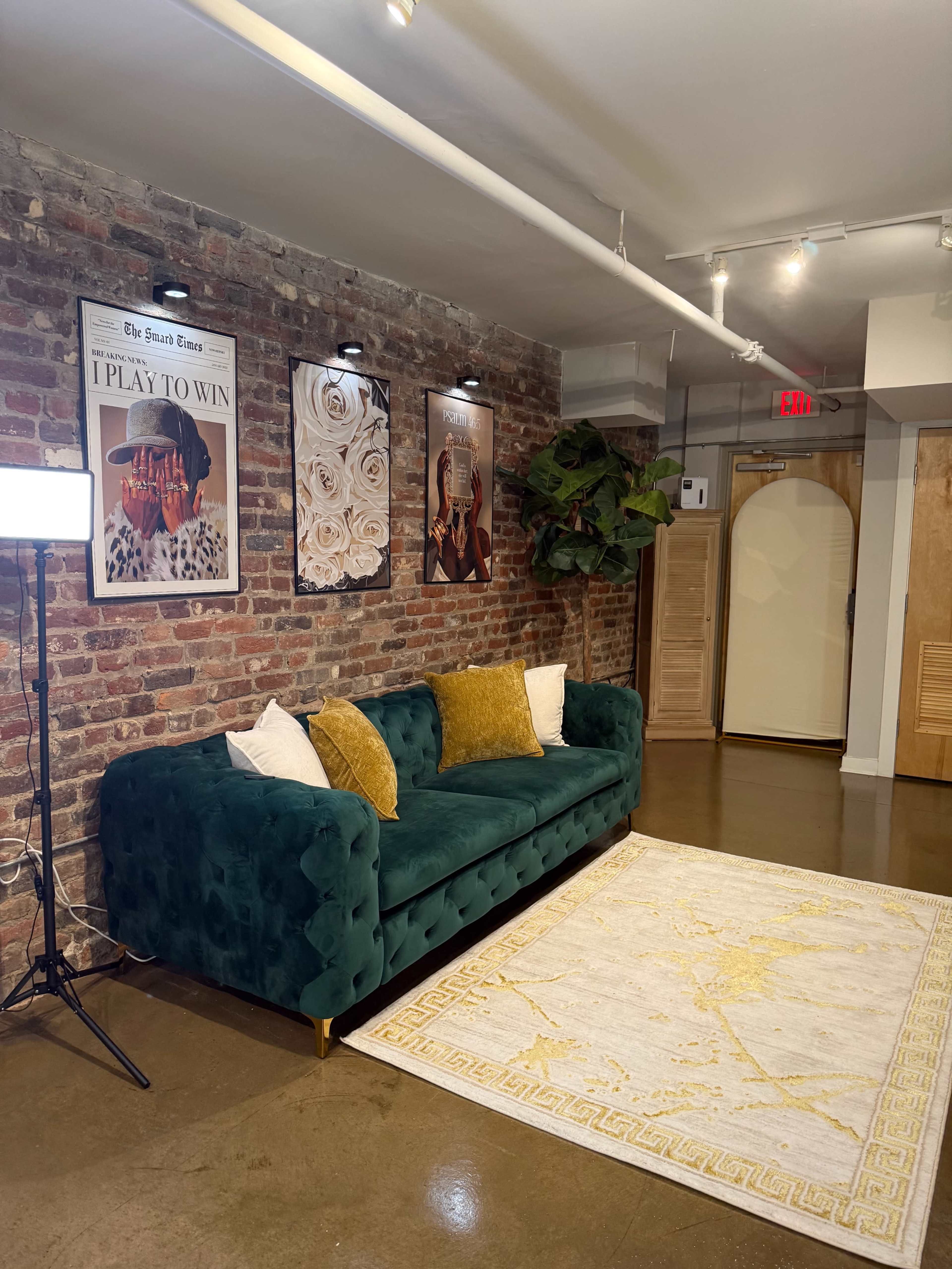 The image shows a green velvet sofa with decorative pillows, positioned against a brick wall adorned with framed artwork, illuminated by overhead lights in a modern interior space.