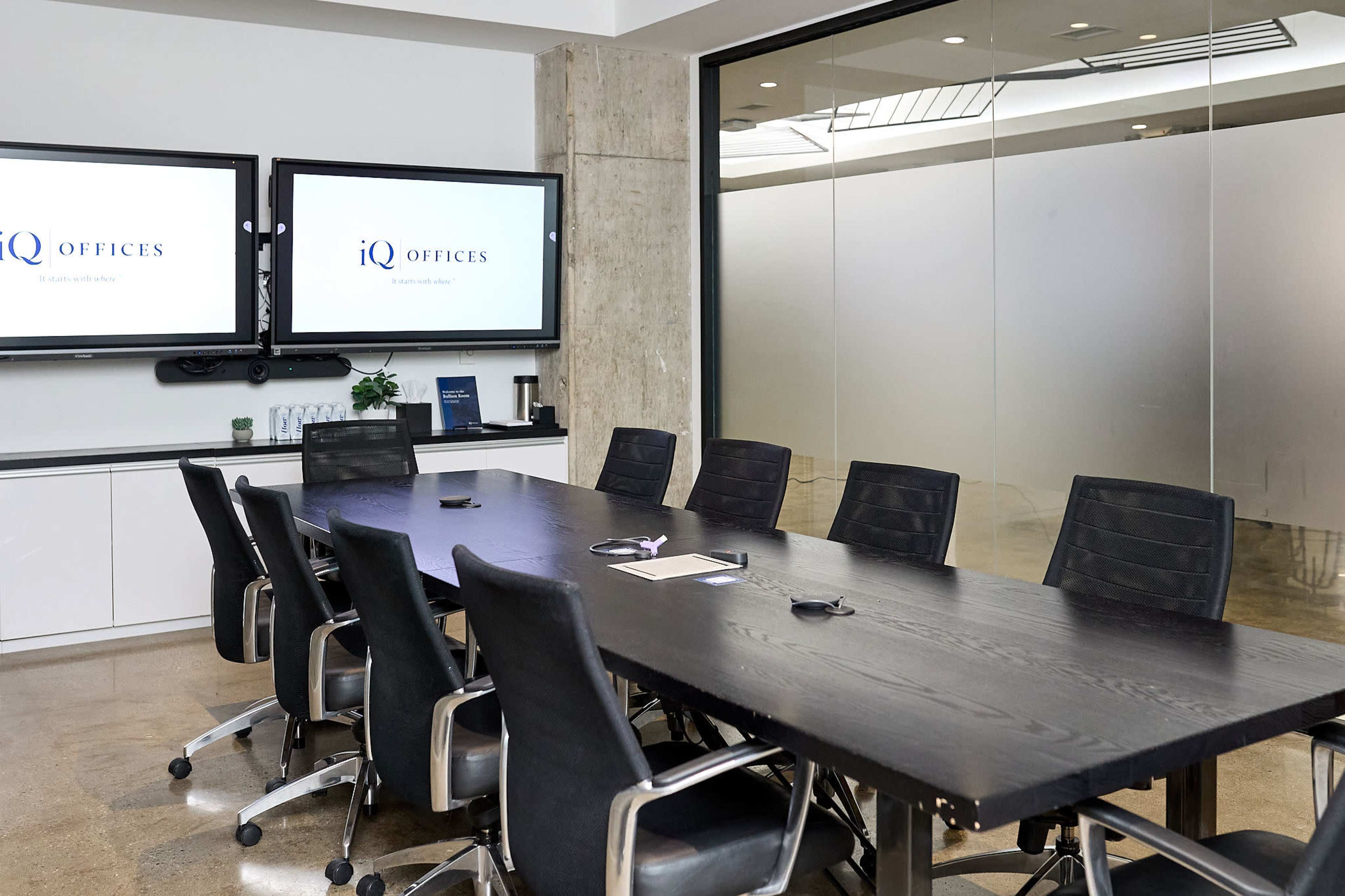 The image shows a modern conference room featuring a large black table surrounded by ergonomic chairs, two wall-mounted screens displaying the logo "iQ Offices," and a glass partition reflecting the room's interior.