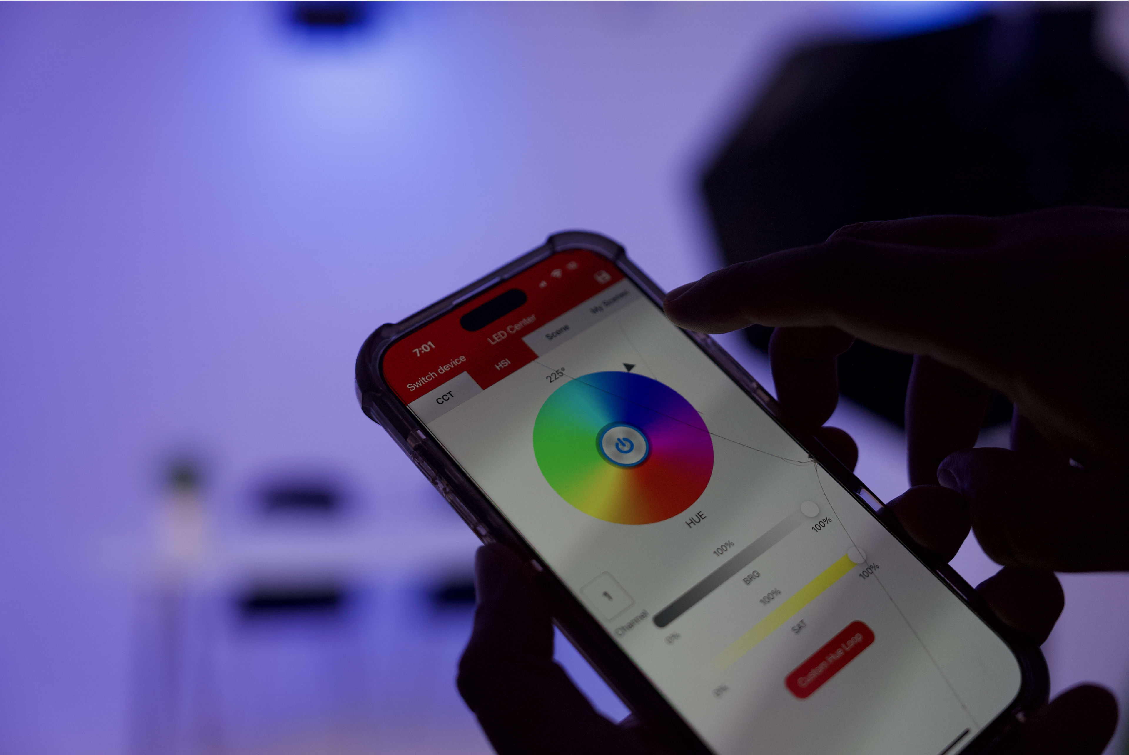 A person is using a smartphone app to adjust LED color settings, displaying a color wheel and sliders on the screen.