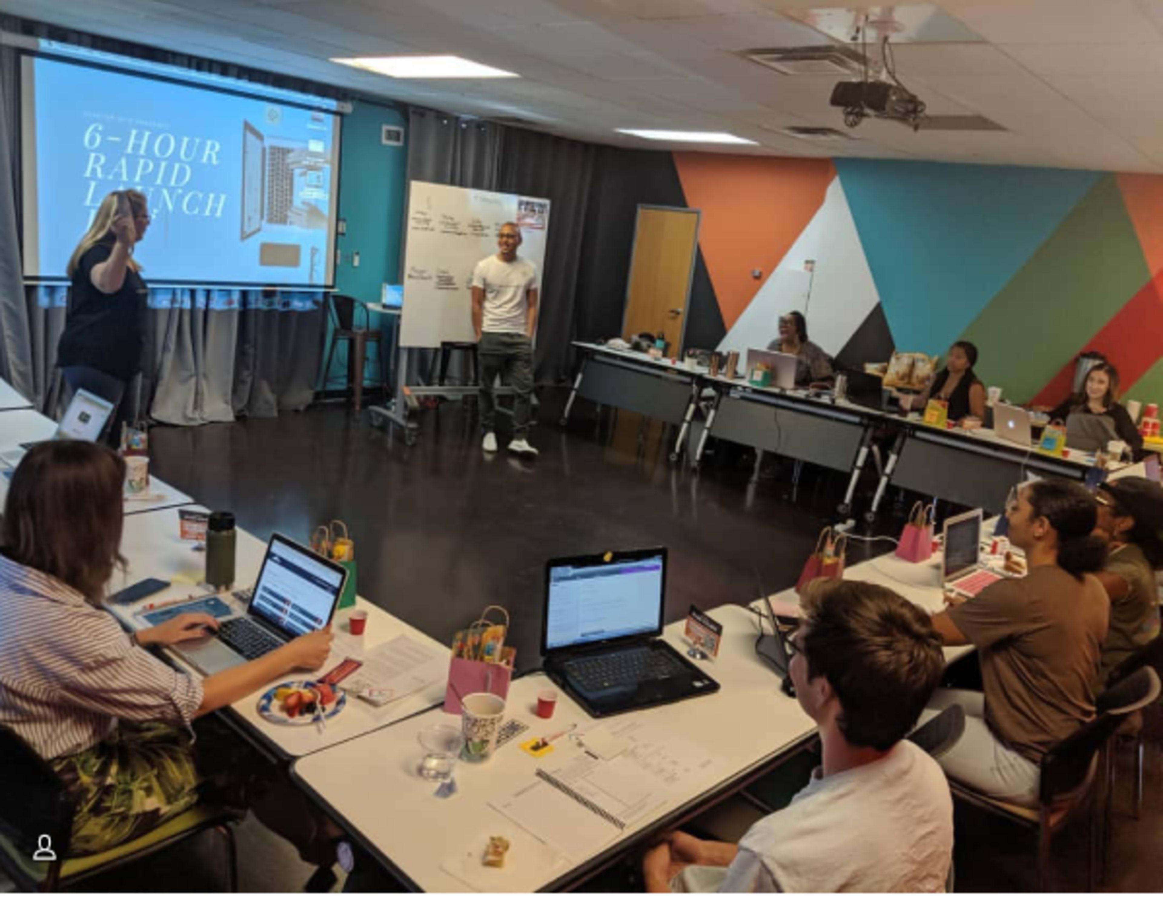 A group of people are seated at tables in a classroom setting, actively participating in a workshop titled "6-Hour Rapid Launch," with a presentation visible on a screen in the background.
